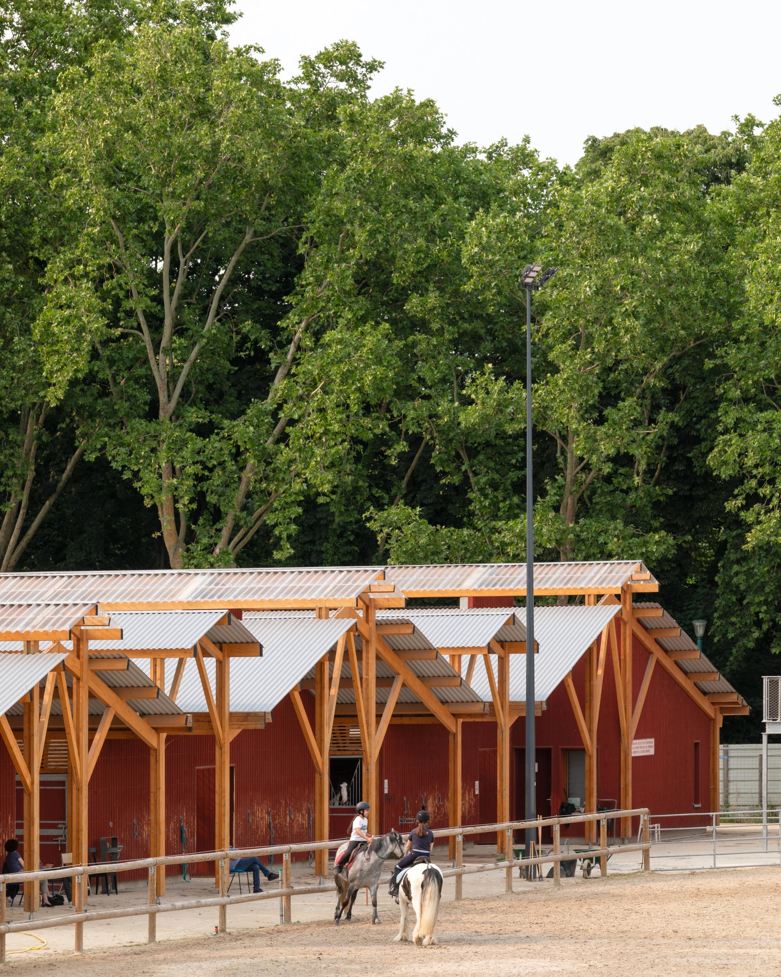 Centre équestre de Gennevilliers : écuries en bois bardées de rouge de Falun, toitures légères en sheds métalliques bordant la carrière au pied d’un parc arboré.