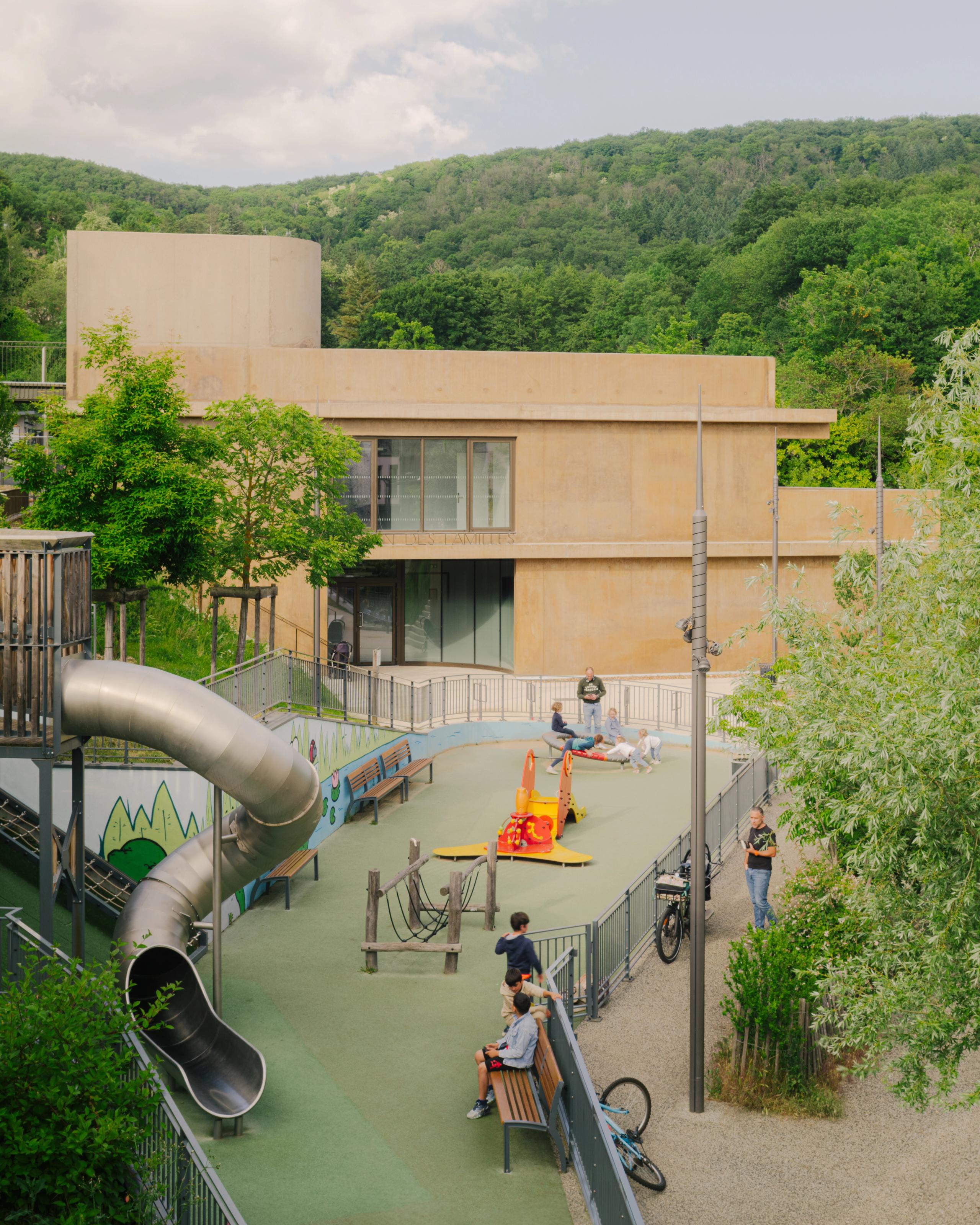 Maison des familles à Limonest : volume en béton teinté en strates, parvis en belvédère sur aire de jeux en pente, intégrée au versant boisé.
