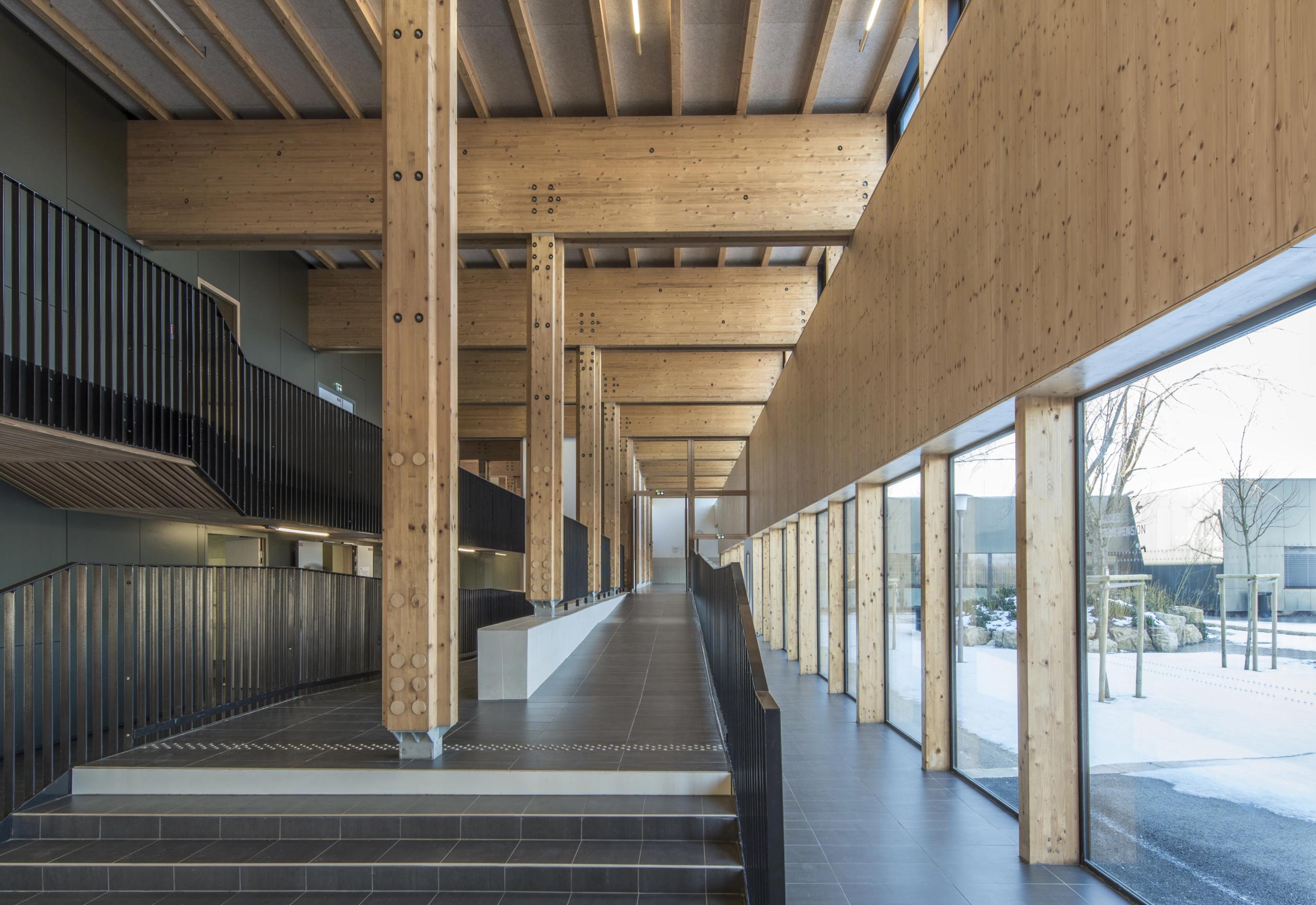 Grande galerie traversante du collège Pierre-Joseph Proudhon à Besançon, structure poteau-poutre bois apparente, rampes noires et façade vitrée sur cour enneigée.