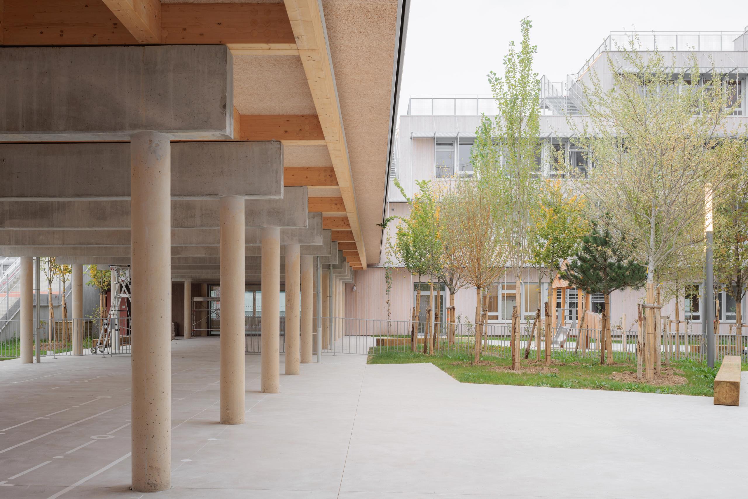 Covered concrete-and-timber walkway framing the courtyard of Dominique Frelaut School Complex in Colombes, opening onto planted garden and façades.