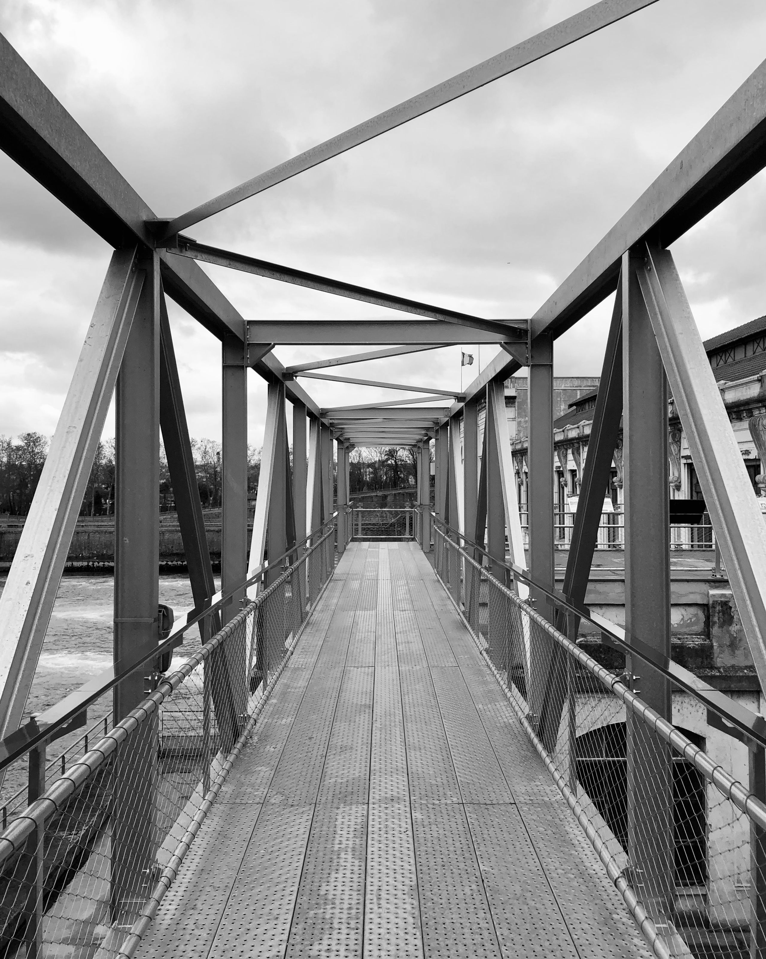 Passerelle métallique du poste source de Cusset à Villeurbanne, structure en treillis galvanisé cadrant la perspective et les circulations au-dessus de l’eau.