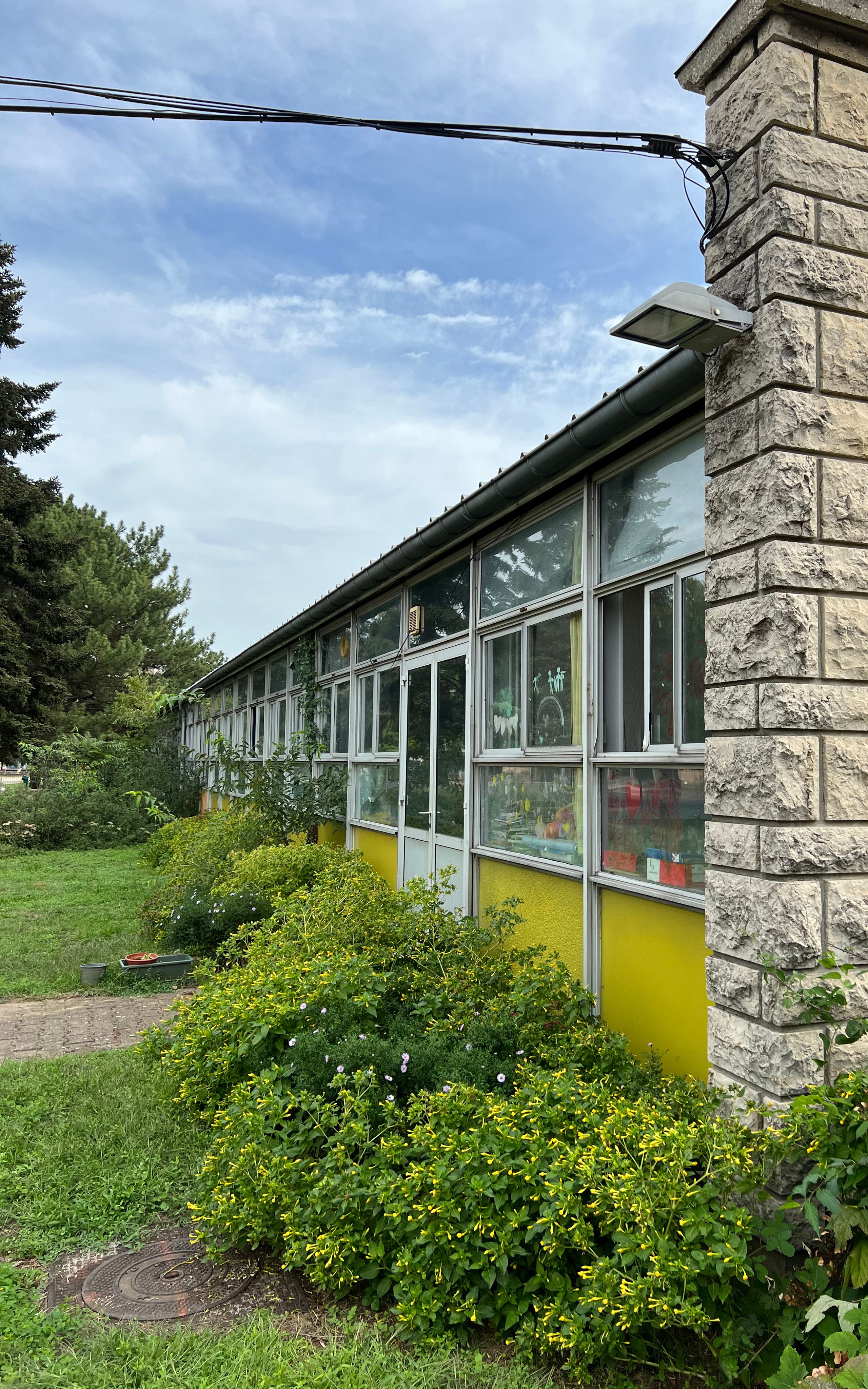 Façade existante du groupe scolaire Kennedy à Lyon : long volume vitré à allège jaune, structure légère et jardin dense en premier plan.