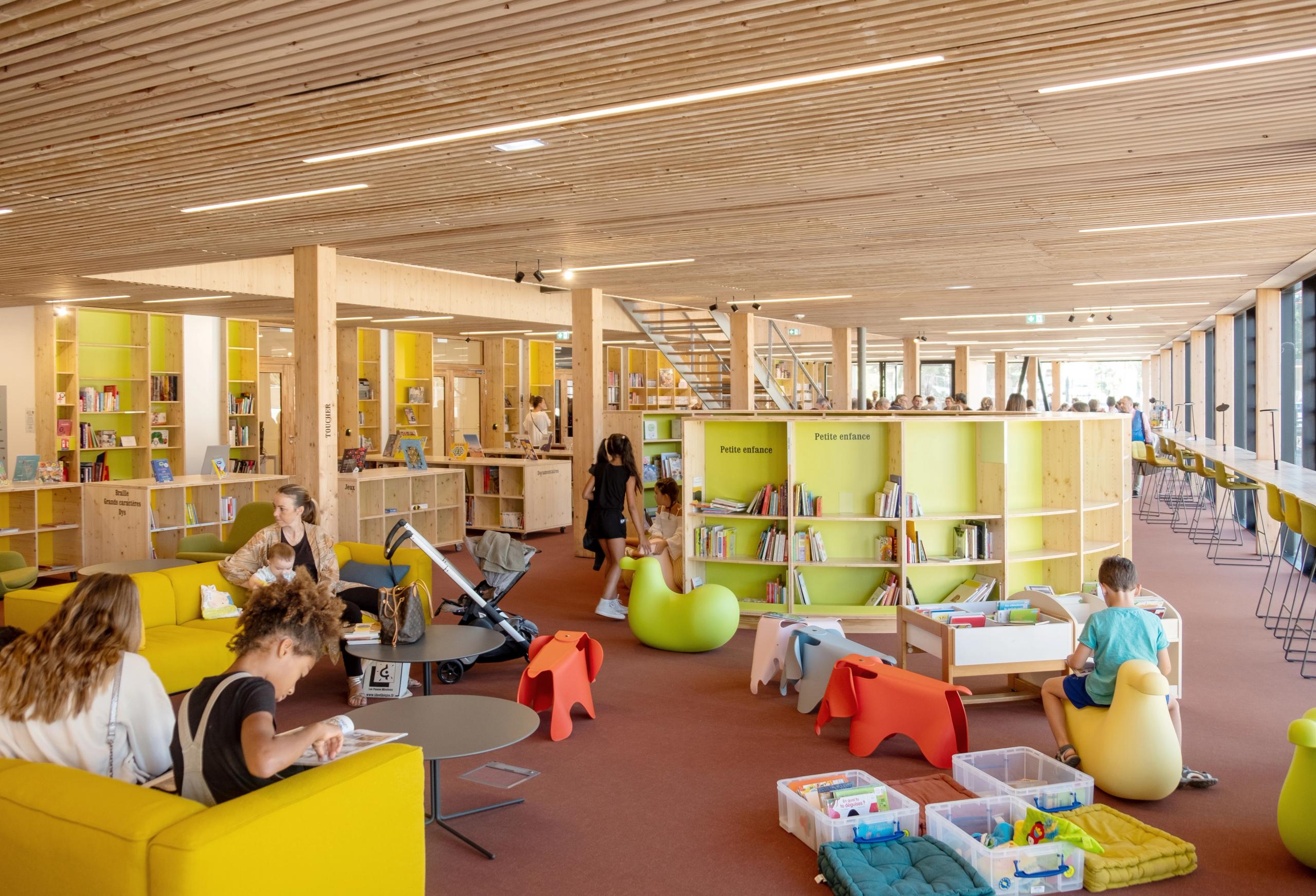 Intérieur en bois de la médiathèque du groupe scolaire Claudie Haigneré aux Pennes-Mirabeau, espace jeunesse ouvert avec mobilier coloré et lumière naturelle.