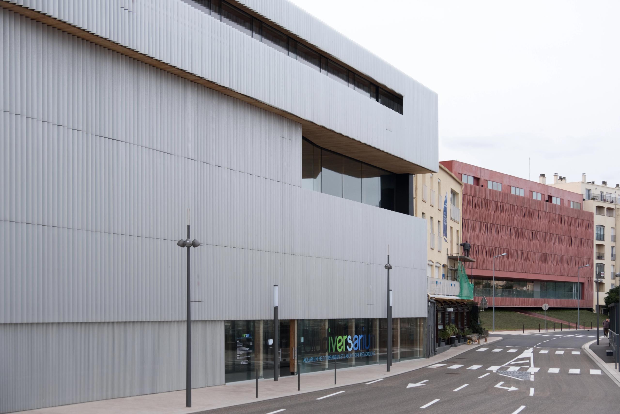 Façade compacte du Biodiversarium à Banyuls-sur-Mer, volume horizontal bardé de métal ondulé microperforé, percé de vitrages en bande sur rue portuaire.