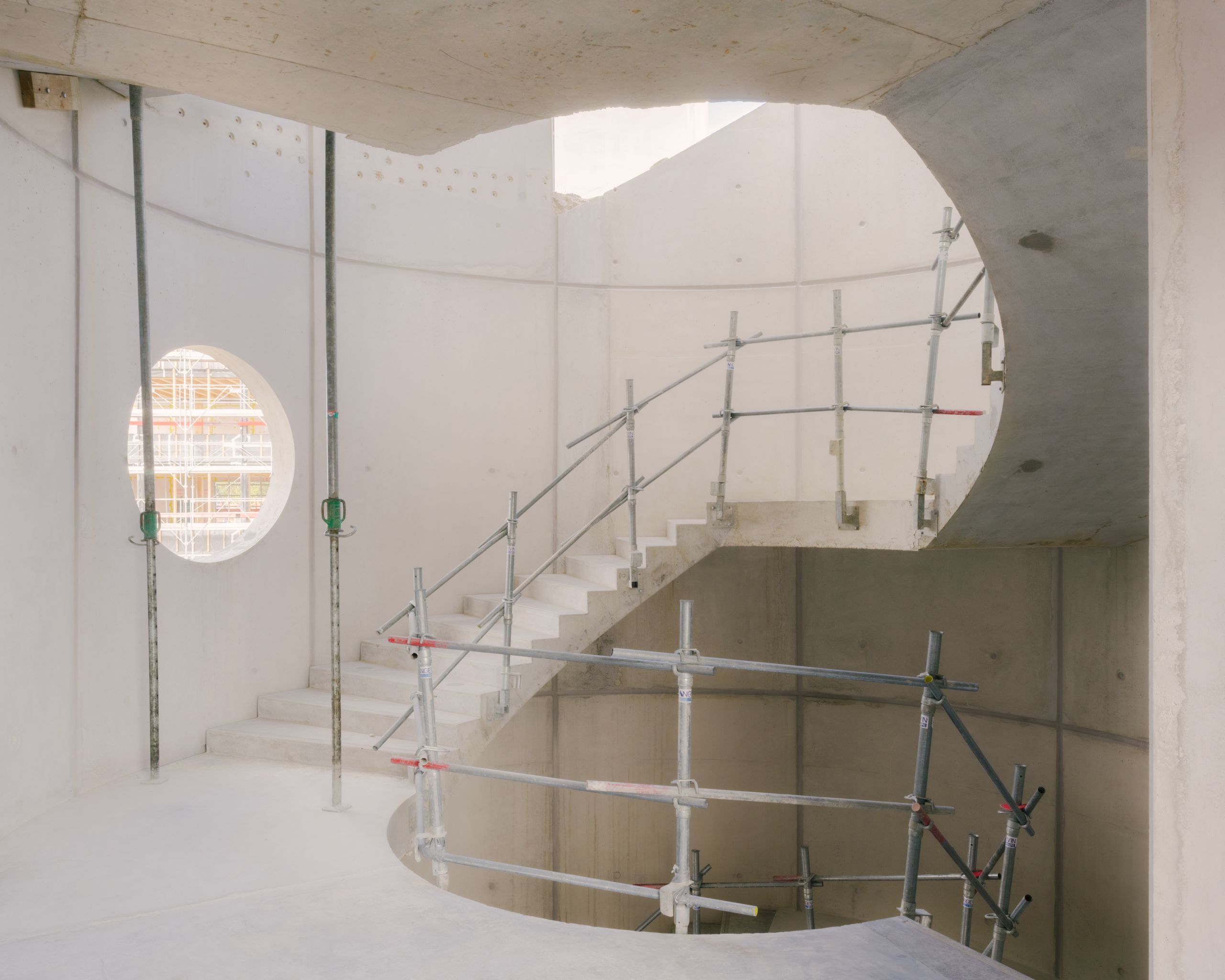 Escalier en béton courbe en chantier du Groupe scolaire Dominique Frelaut à Colombes, noyau central éclairé par un oculus et ceinturé d’échafaudages métalliques.