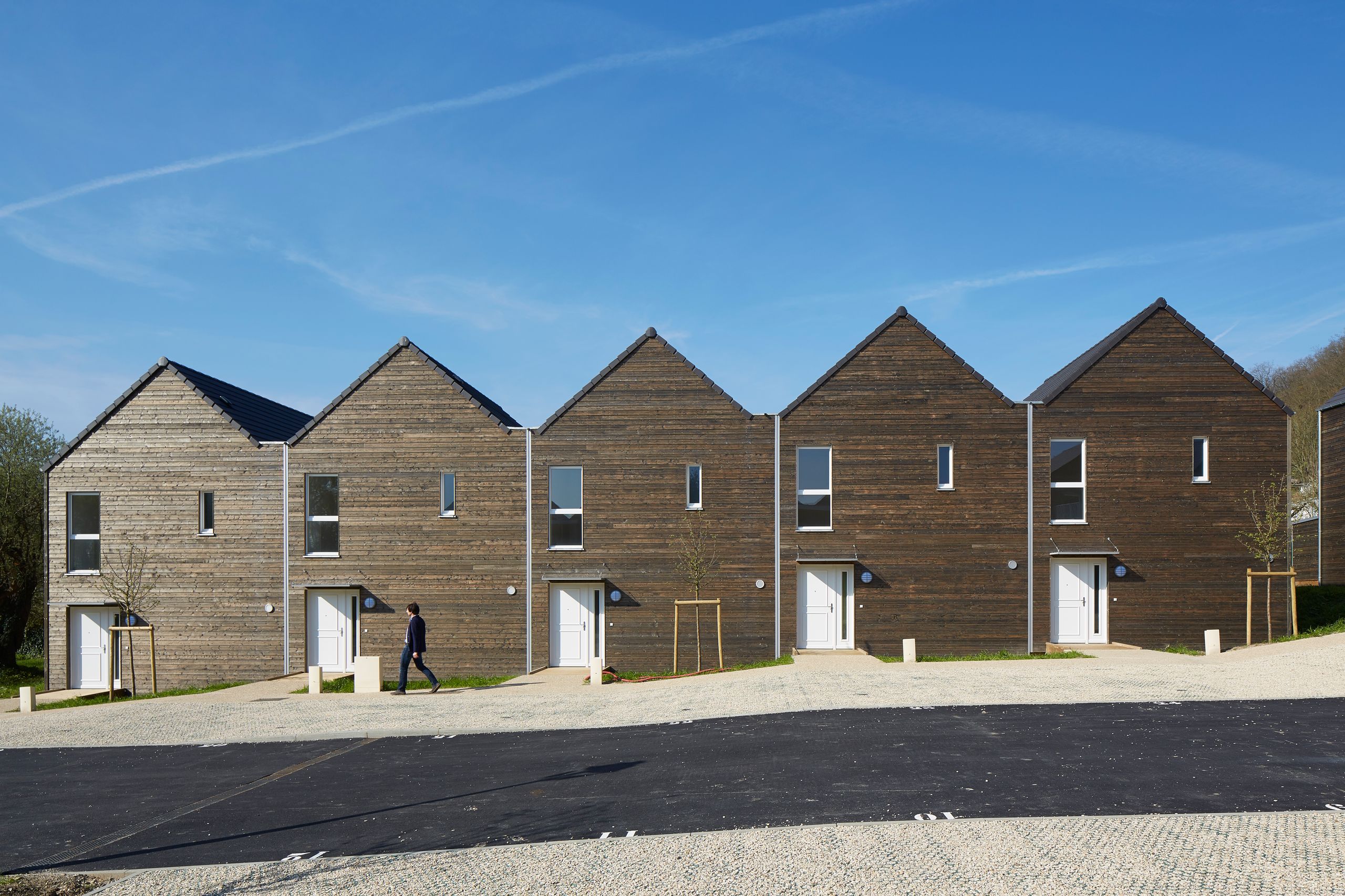 Façade alignée des 36 logements Tectoniques à Vauhallan, volumes compacts en bois à toitures à deux pans, percements verticaux et accès individuels sur espace minéral.