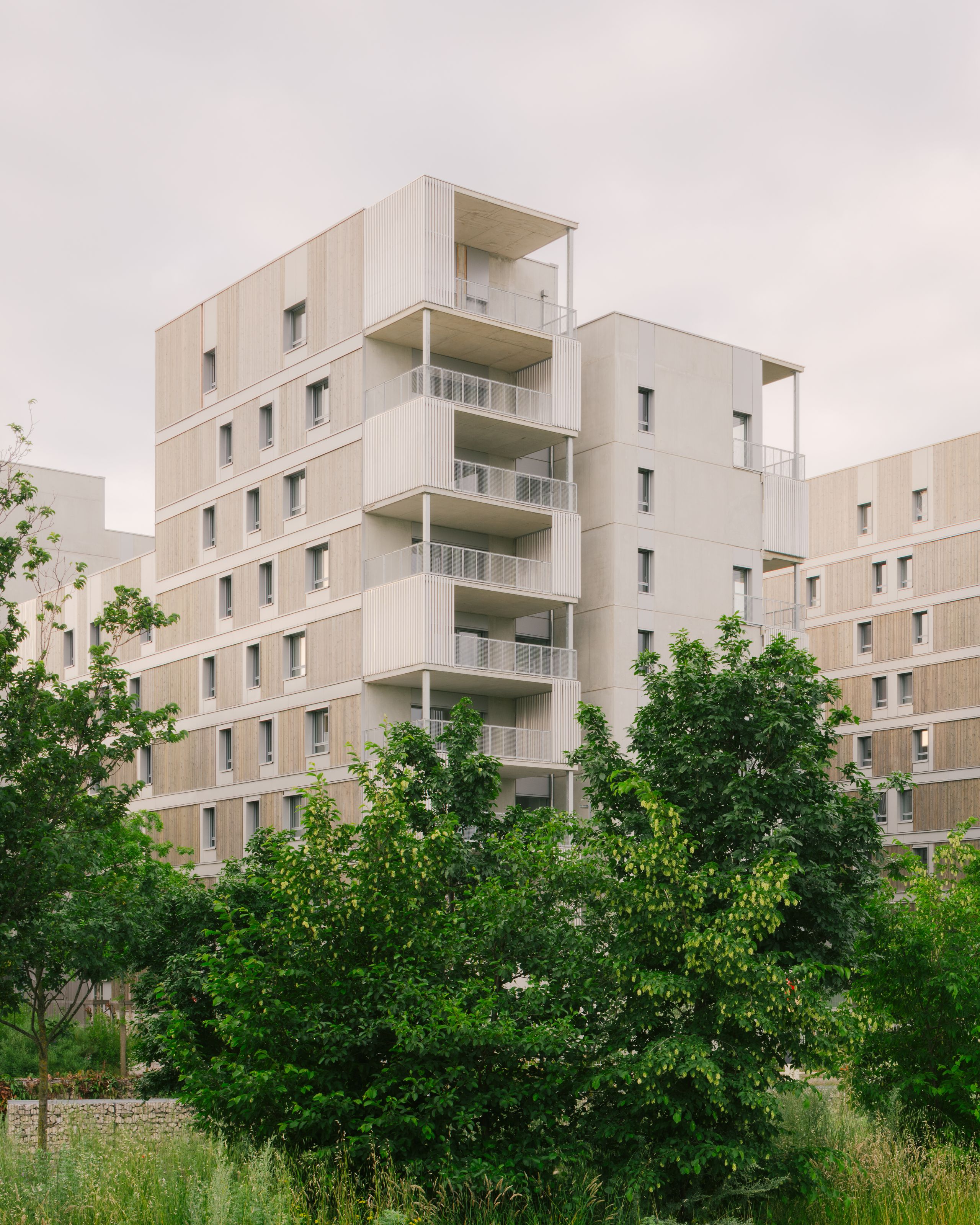 Vue du projet de 116 logements à Vénissieux : volumes fragmentés en R+6, façades bois-béton, balcons en exosquelette métallique, en lisière d’un jardin planté.