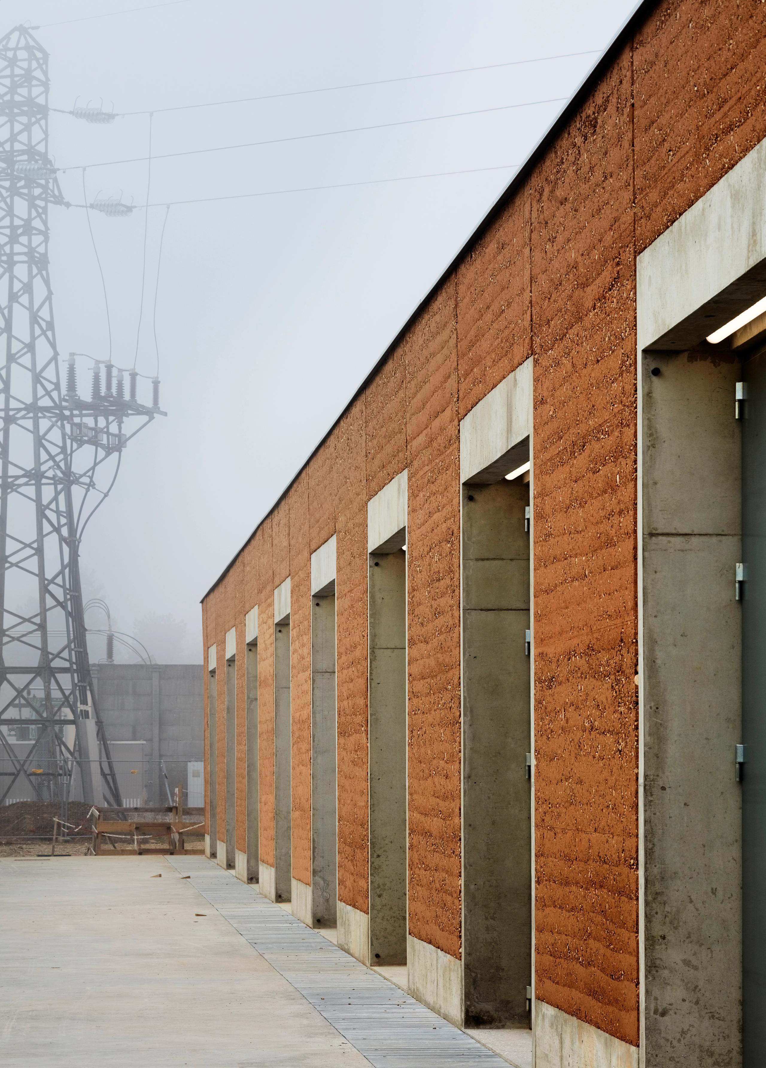 Façade en pisé et béton brut du poste source de Cusset à Villeurbanne, rythmée par une série de baies verticales, dans un paysage industriel brumeux.