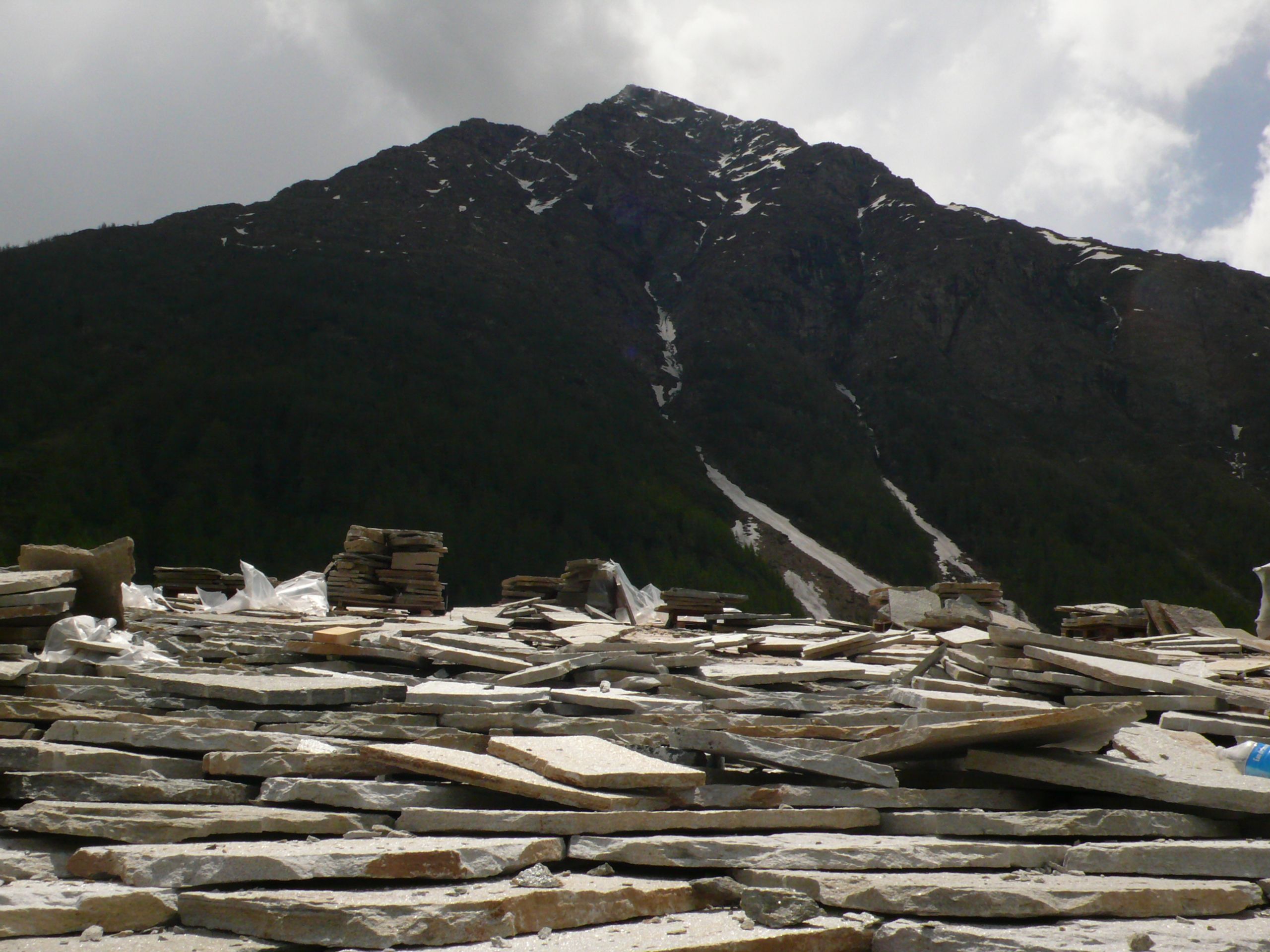 Stock de lauzes grises à ciel ouvert pour l’Espace nordique de Bessans, empilées au pied d’un versant montagneux enneigé, en contexte de chantier.