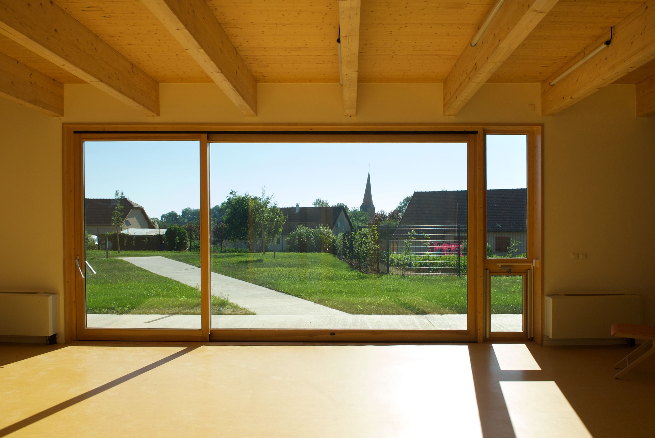 Intérieur en structure bois de l’EHPAD de Mervans, grande baie vitrée ouvrant la salle commune sur le jardin clos et le paysage pavillonnaire du village.