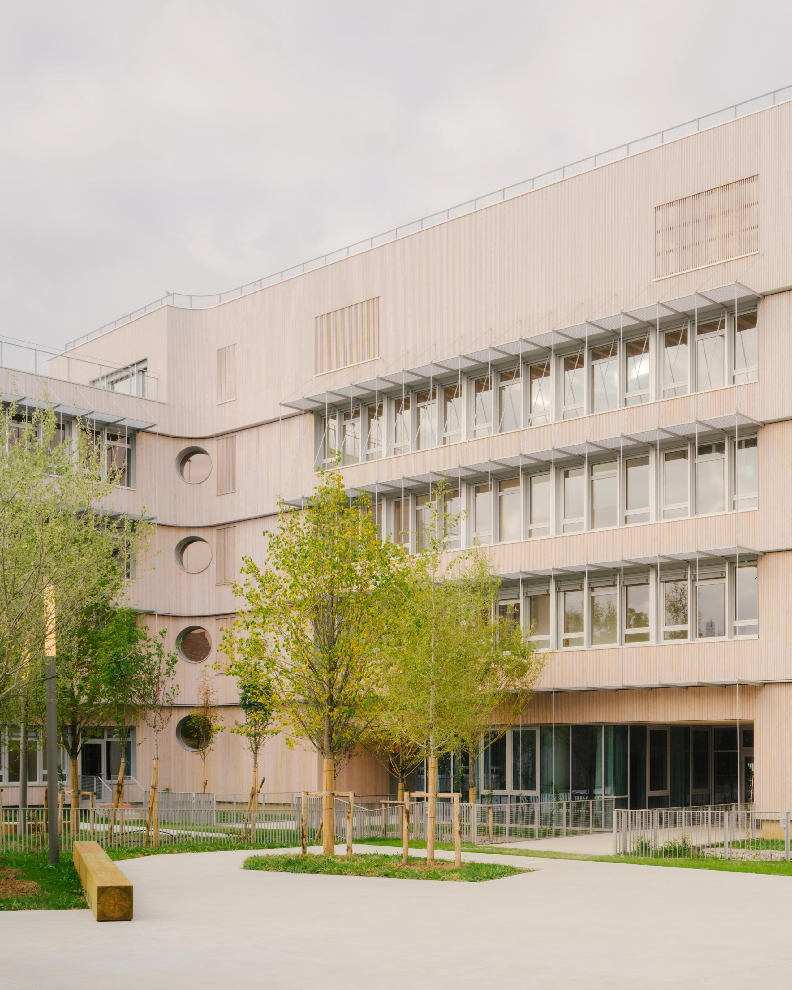 Façade bois du groupe scolaire Dominique Frelaut à Colombes, volumes en U sur cour plantée, percements circulaires et casquette métallique filtrant la lumière.