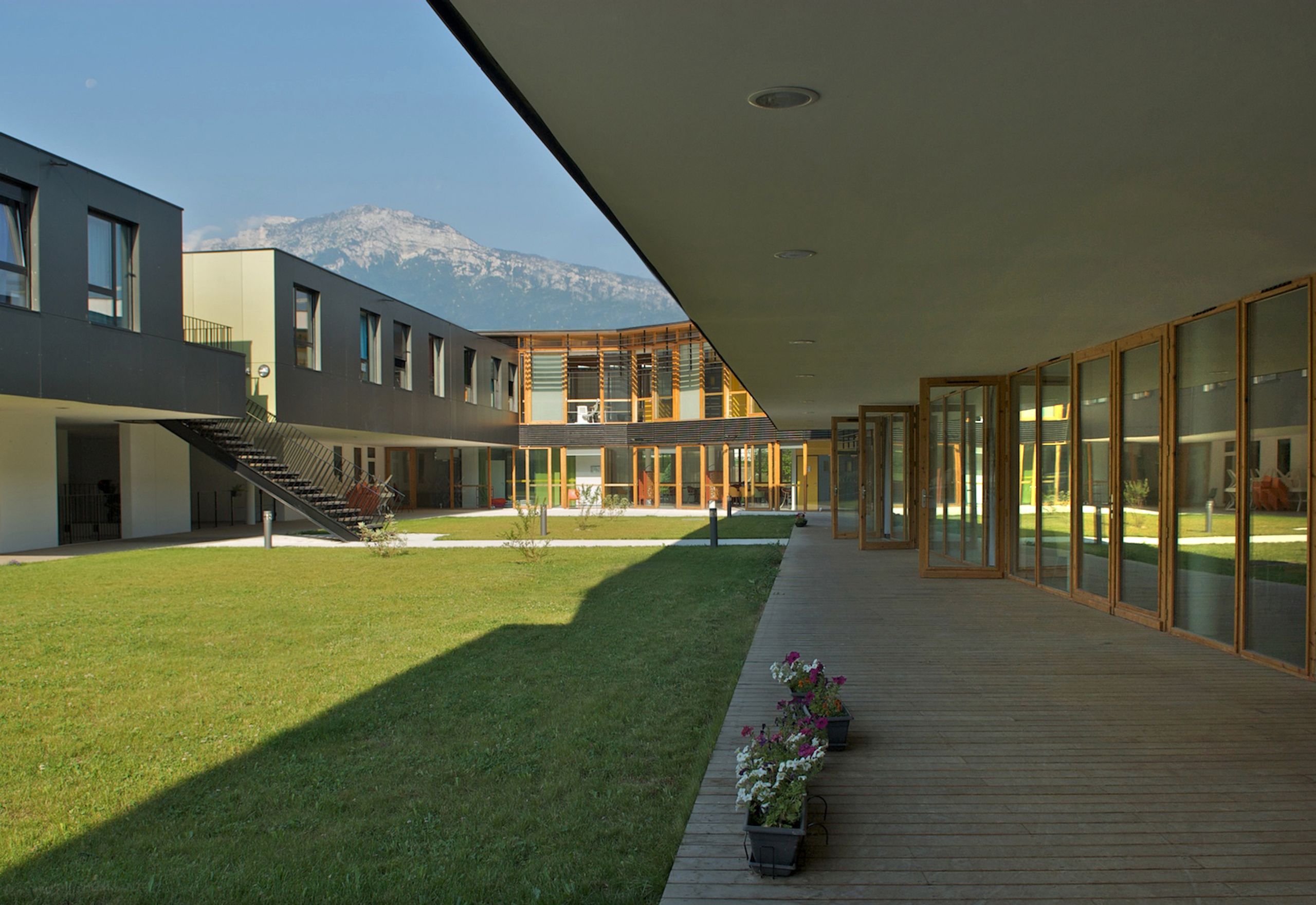 Cour intérieure de l’EHPAD à Grenoble, ailes en U à deux niveaux, larges baies vitrées et résille bois, circulations couvertes ouvertes sur jardin et montagne.