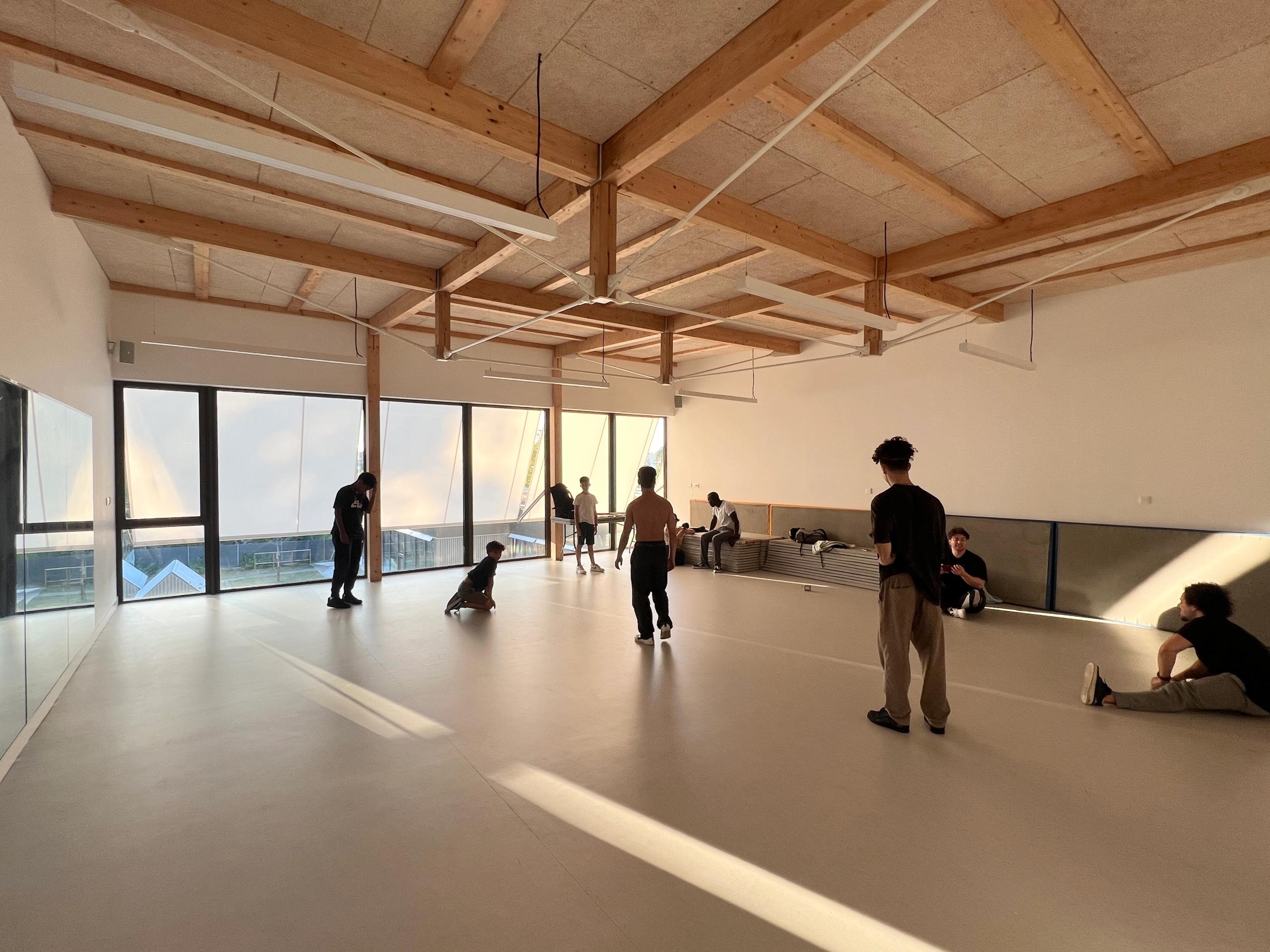 Salle polyvalente de la maison des habitants à Mérignac, structure bois apparente, grands vitrages sur paysage urbain, lumière naturelle traversante.