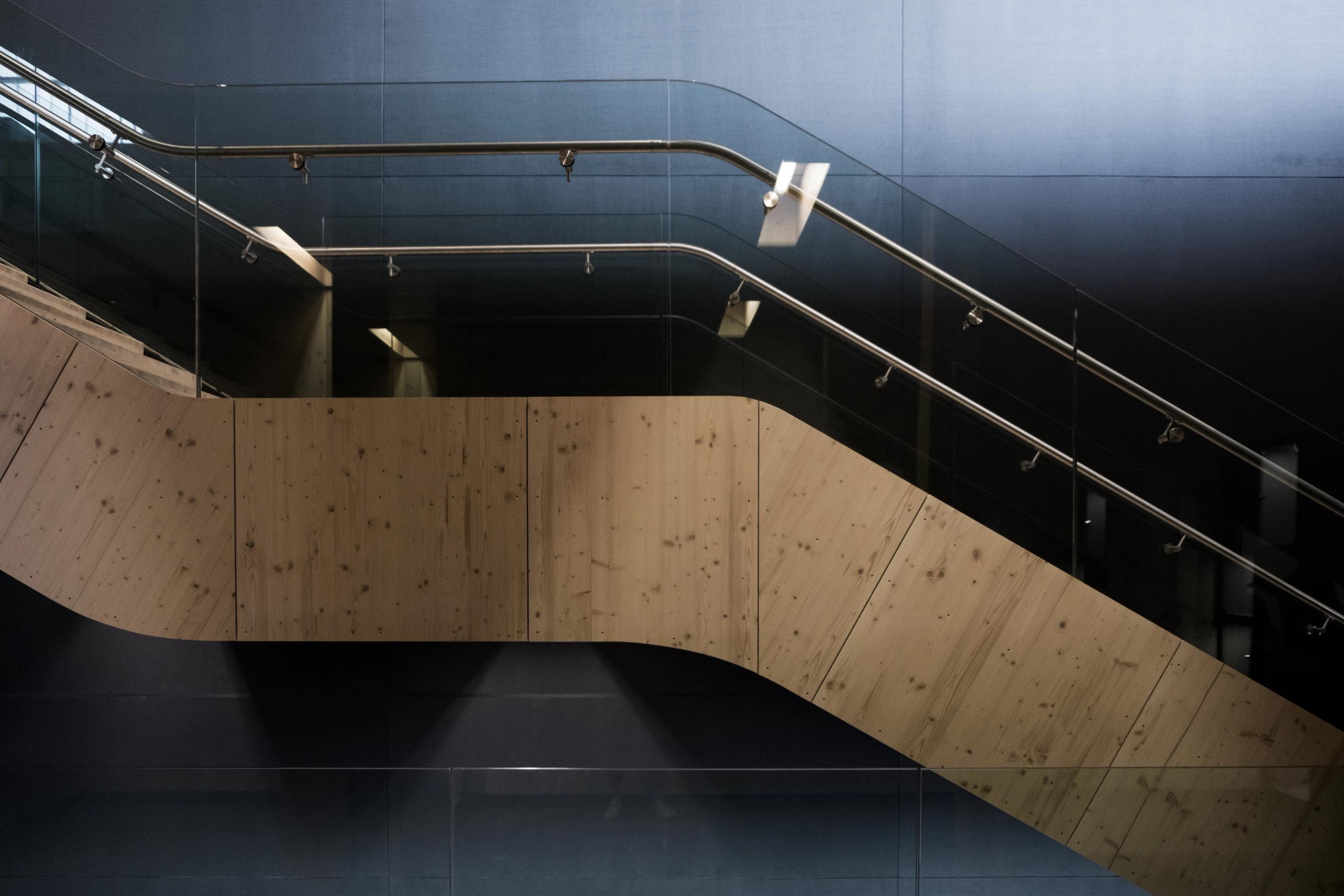 Escalier intérieur du Biodiversarium à Banyuls-sur-Mer, garde-corps vitrés et main courante métallique, noyau en bois clair contrastant avec les parois sombres.
