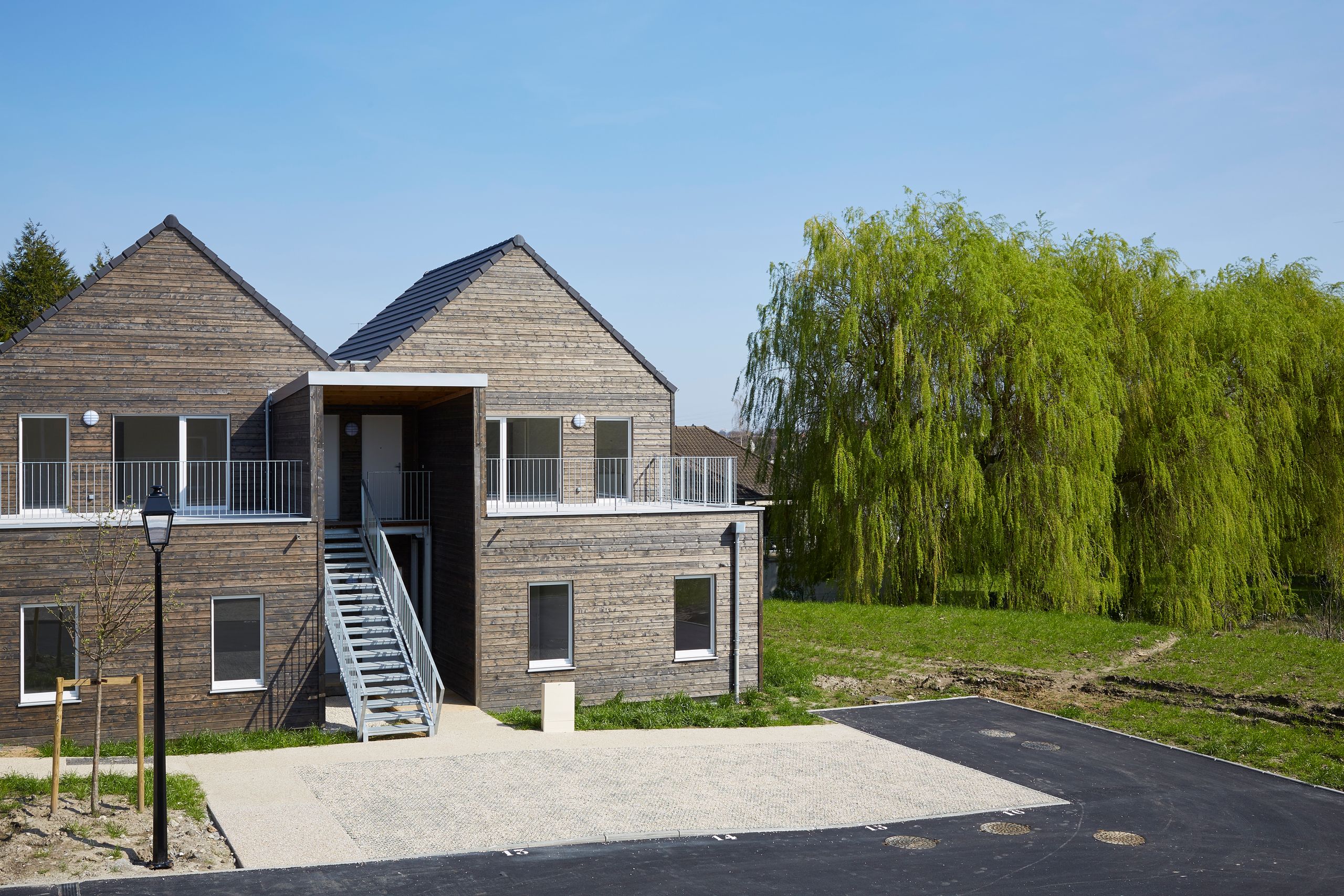 Vue de livraison des 36 logements à Vauhallan par Tectoniques, volumes compacts en bardage bois, toiture à deux pans, coursive et escalier métalliques sur parvis minéral.