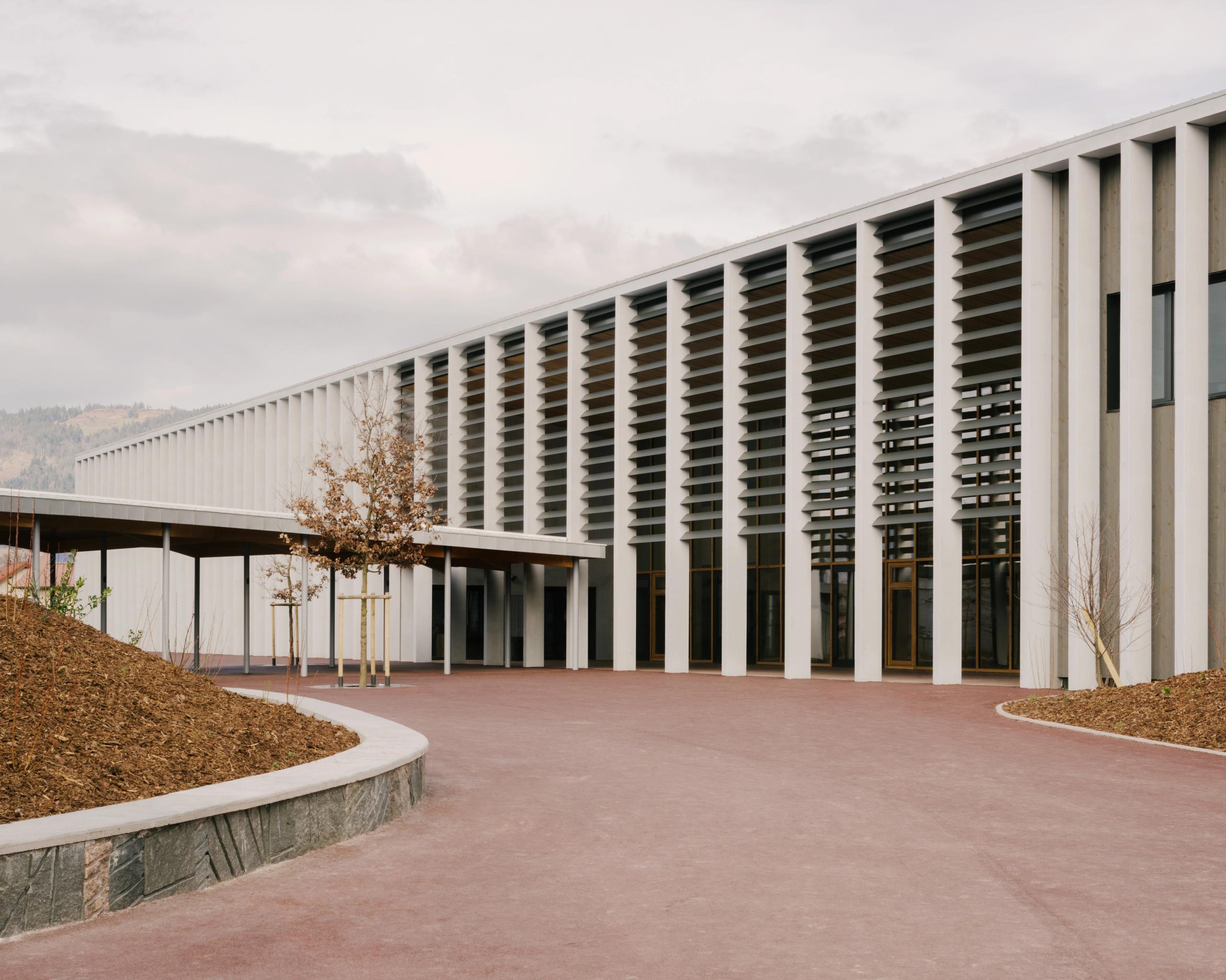 Façade du Collège du Ban à Vagney : portique longitudinal en béton, brise-soleil horizontaux, préau couvert et parvis minéral encadré de talus plantés.