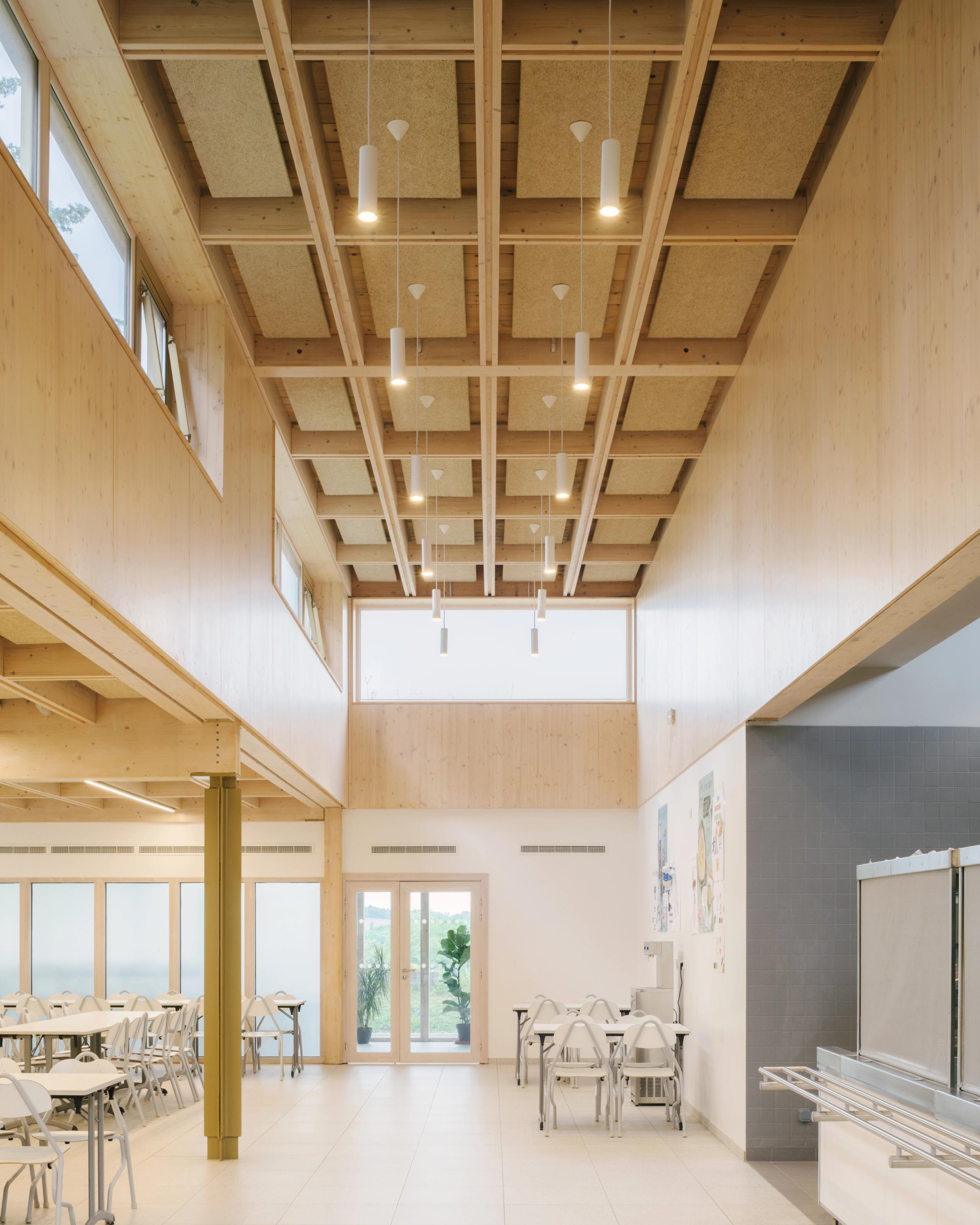 Réfectoire du collège Maryse Bastié à Dole, double hauteur en bois clair, plafond caisson, grandes baies vitrées et mobilier sobre, lumière naturelle abondante.