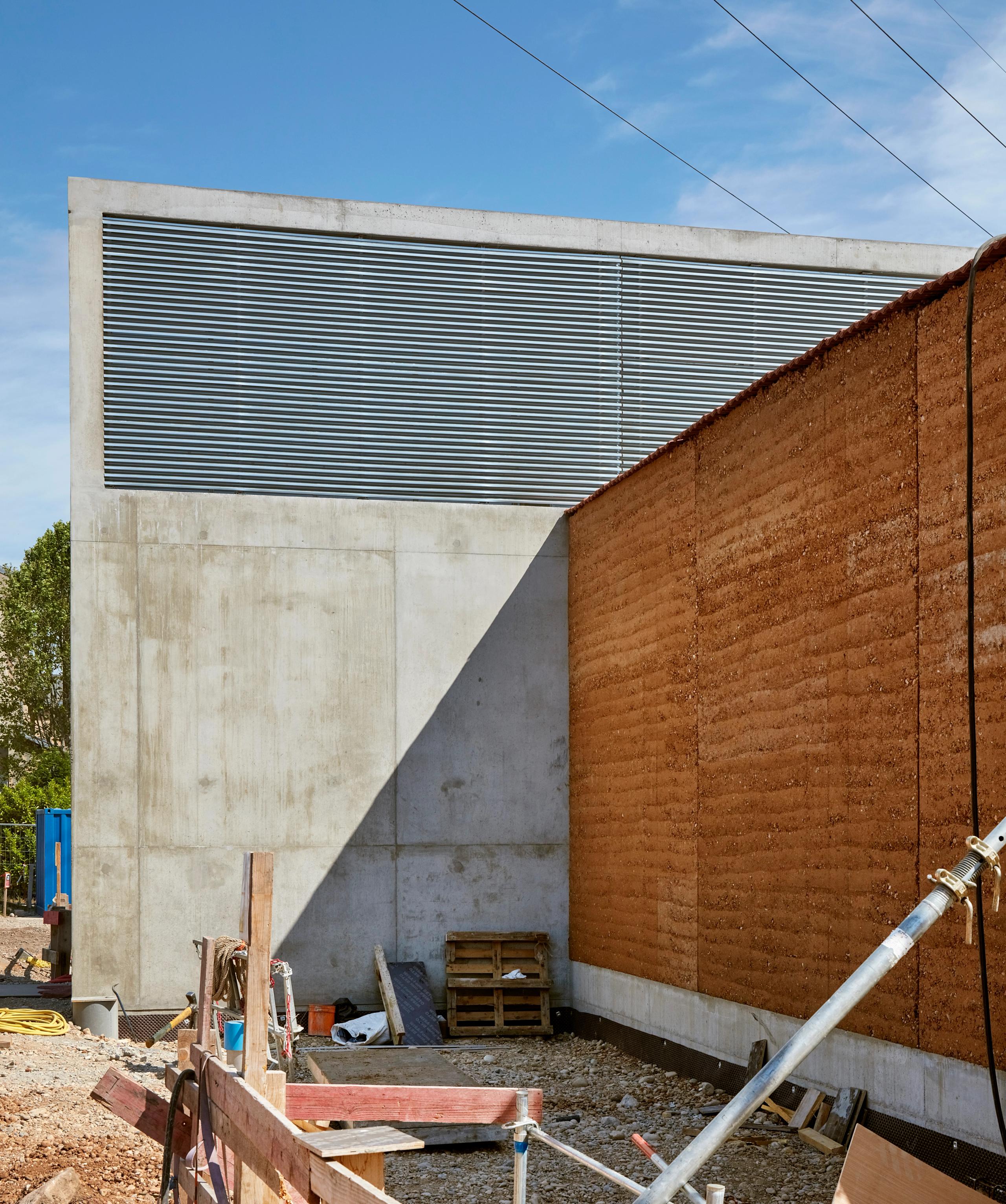 Chantier du poste source de Cusset à Villeurbanne : contraste entre voile en béton brut, mur en pisé et lames métalliques, cadrant une circulation extérieure.