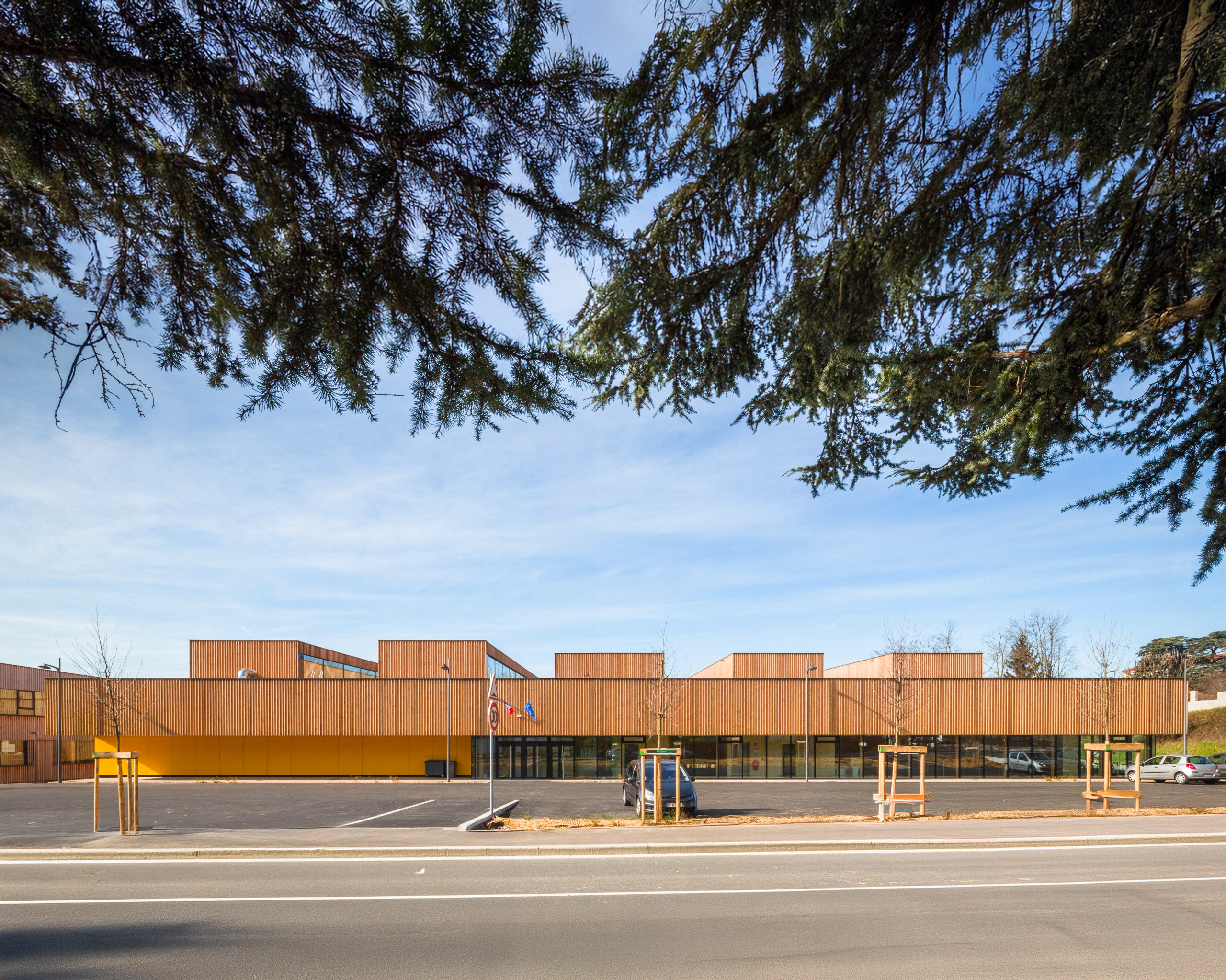 Façade du gymnase Hacine Cherifi à Rillieux-la-Pape, volume bas en bois et socle minéral, sheds nord en toiture, aligné sur une venelle et un parvis minéral.