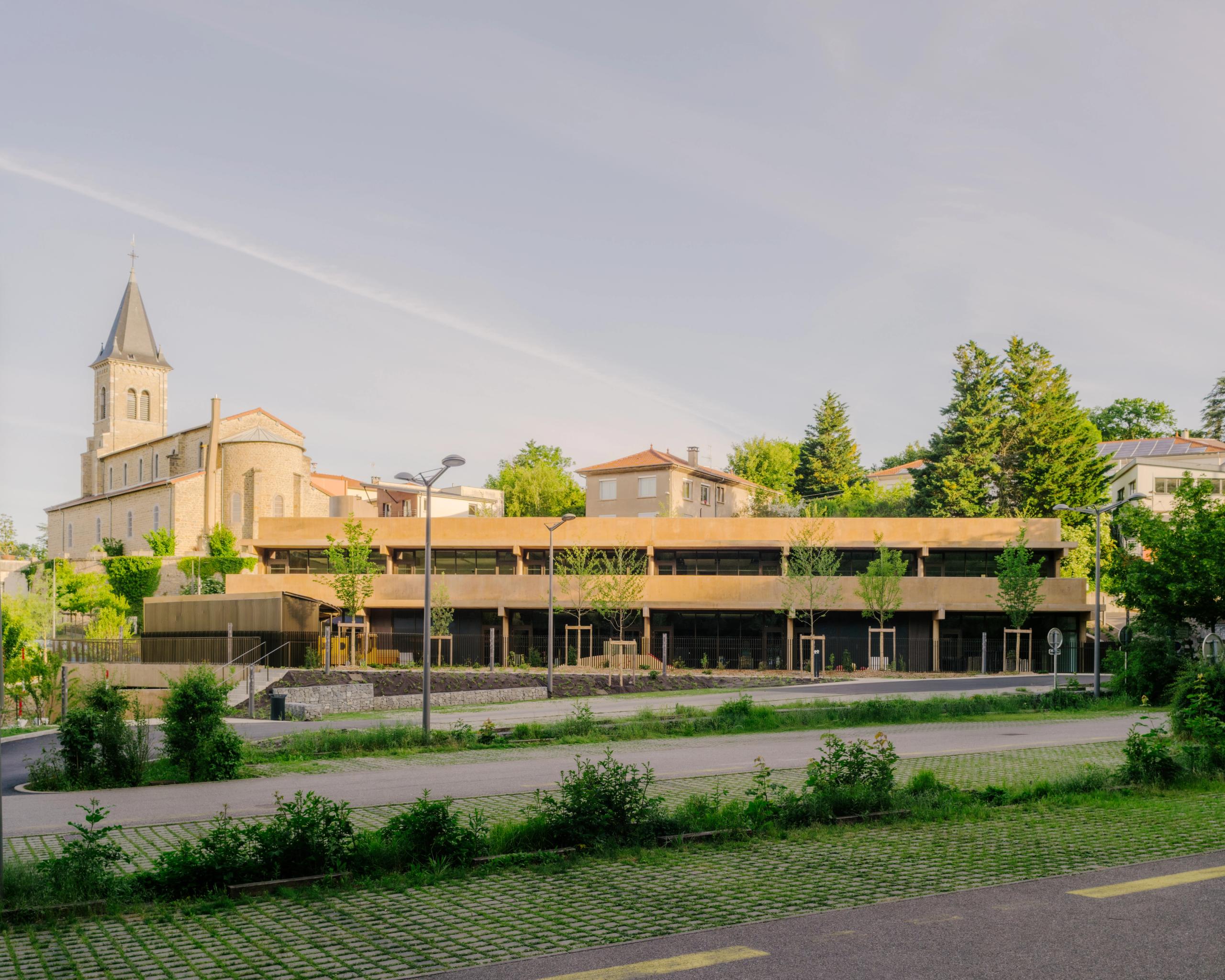 Vue de la Maison des familles à Limonest : volume tellurique en terrasses de béton teinté, façades horizontales vitrées, inséré sous le parvis de l’église.