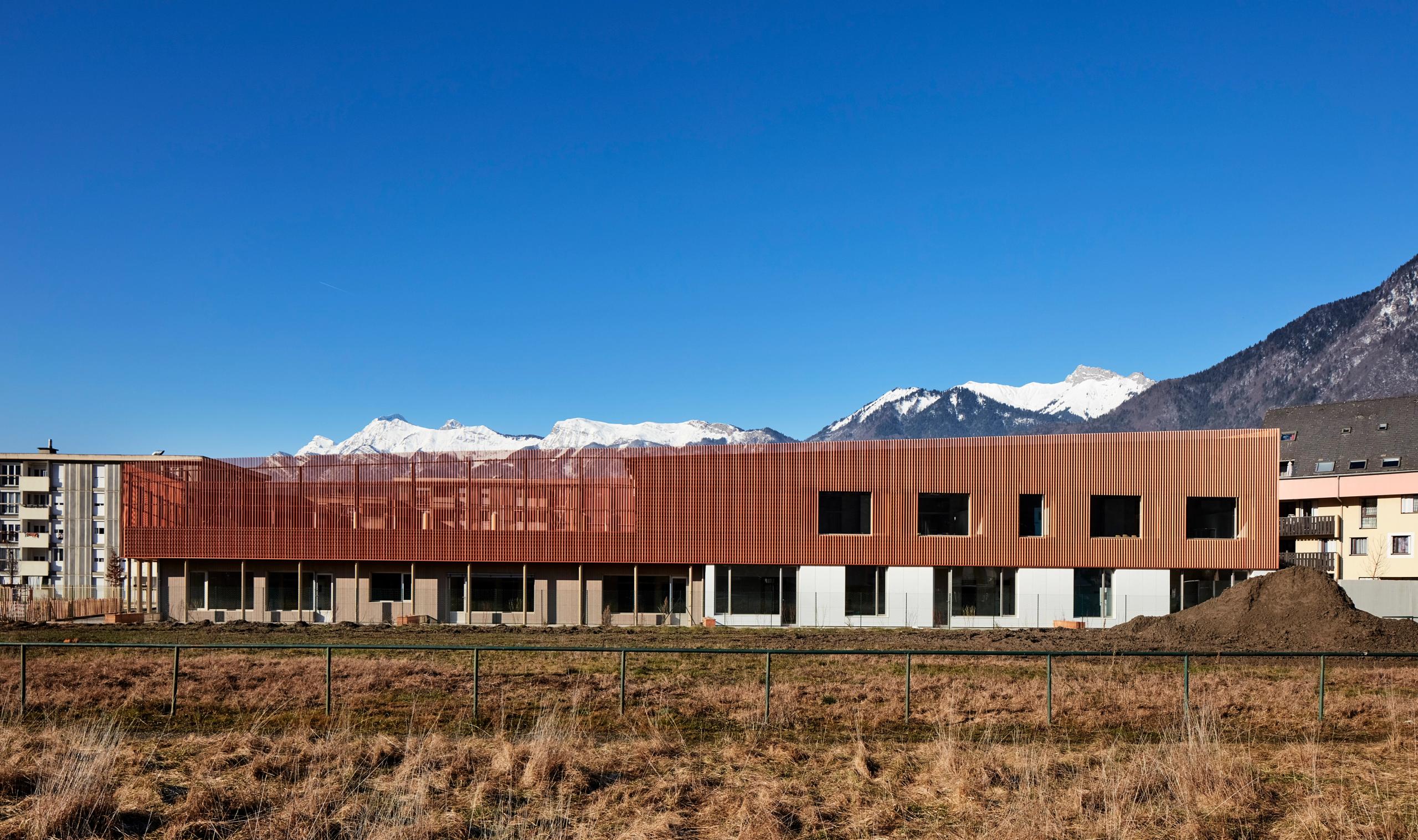 Simone Veil Early Childhood Center in Albertville, a compact copper-toned metal volume with deep window bays, set against snow-covered Alpine mountains.