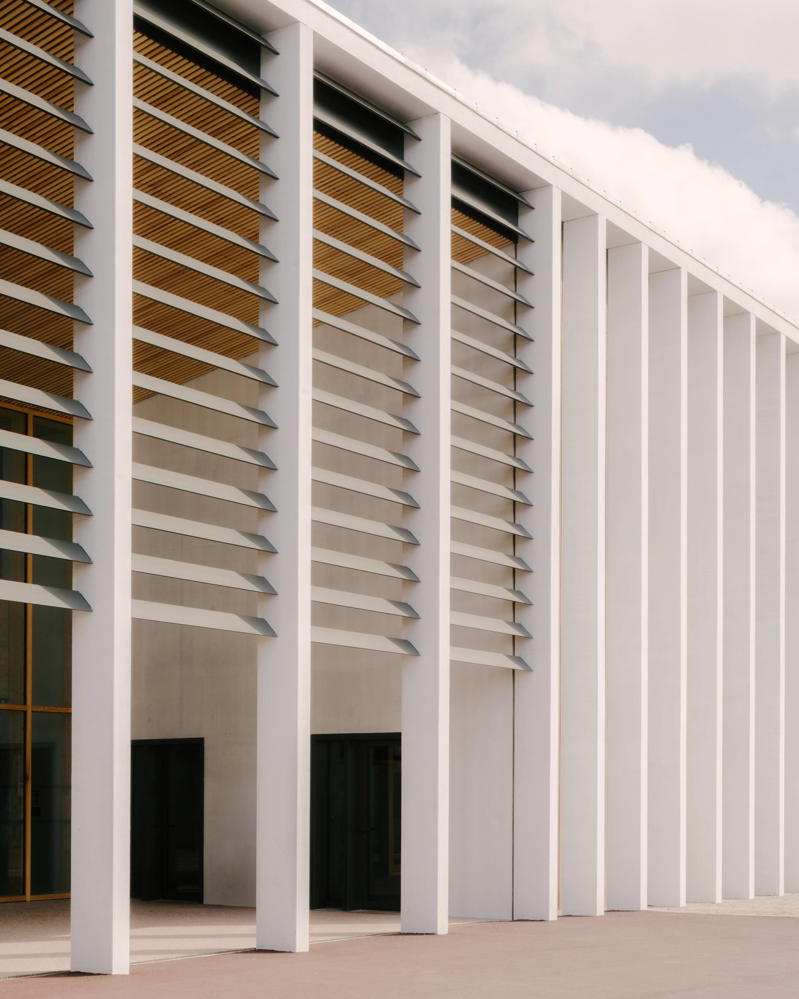 Façade du Collège du Ban à Vagney : portique monumental en béton blanc, lames brise-soleil horizontales, structure rythmique cadrant le préau ouvert.