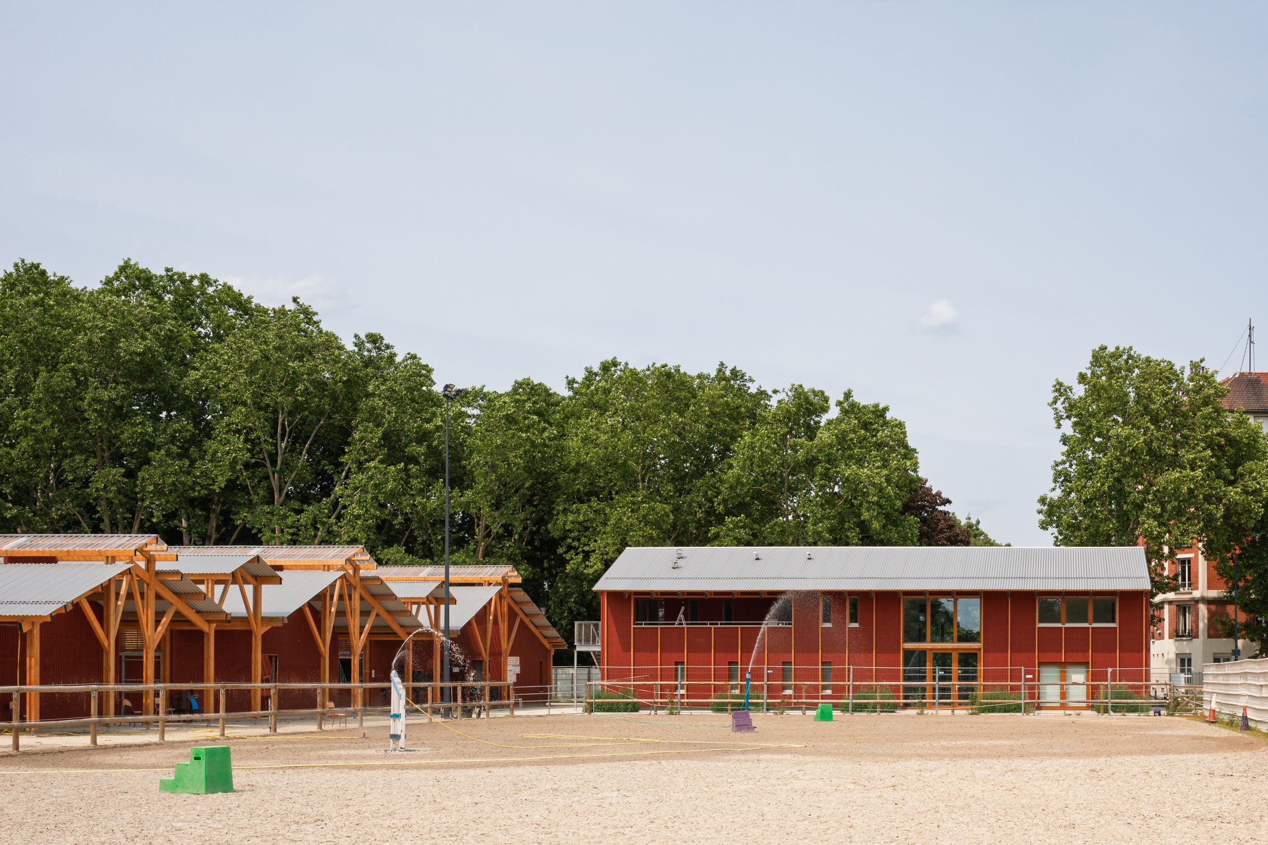 Centre équestre de Gennevilliers : bâtiments en bois bardés de rouge de Falun bordant la carrière, structures apparentes et toitures métalliques face au parc.