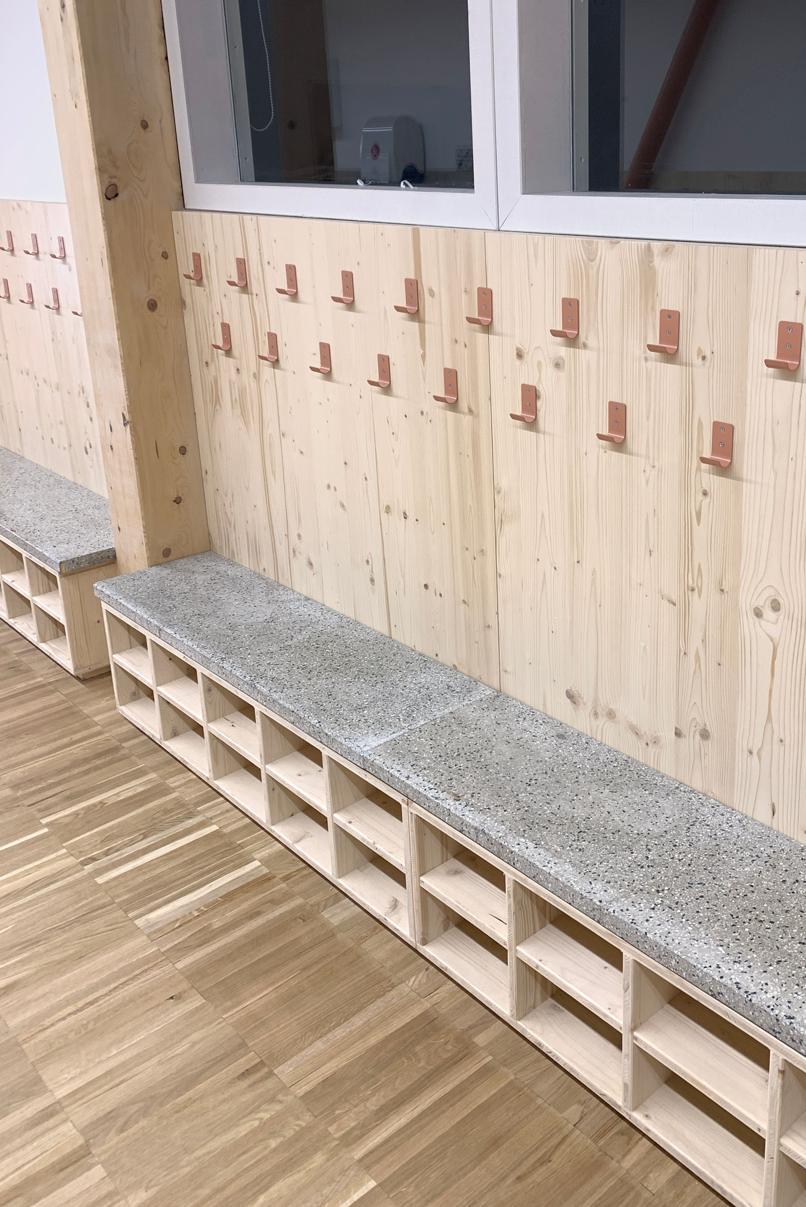 Vestiaire intérieur du Groupe scolaire Kennedy à Lyon, avec banc en béton sur casiers bois, paroi en panneaux bois et alignement de patères métalliques.