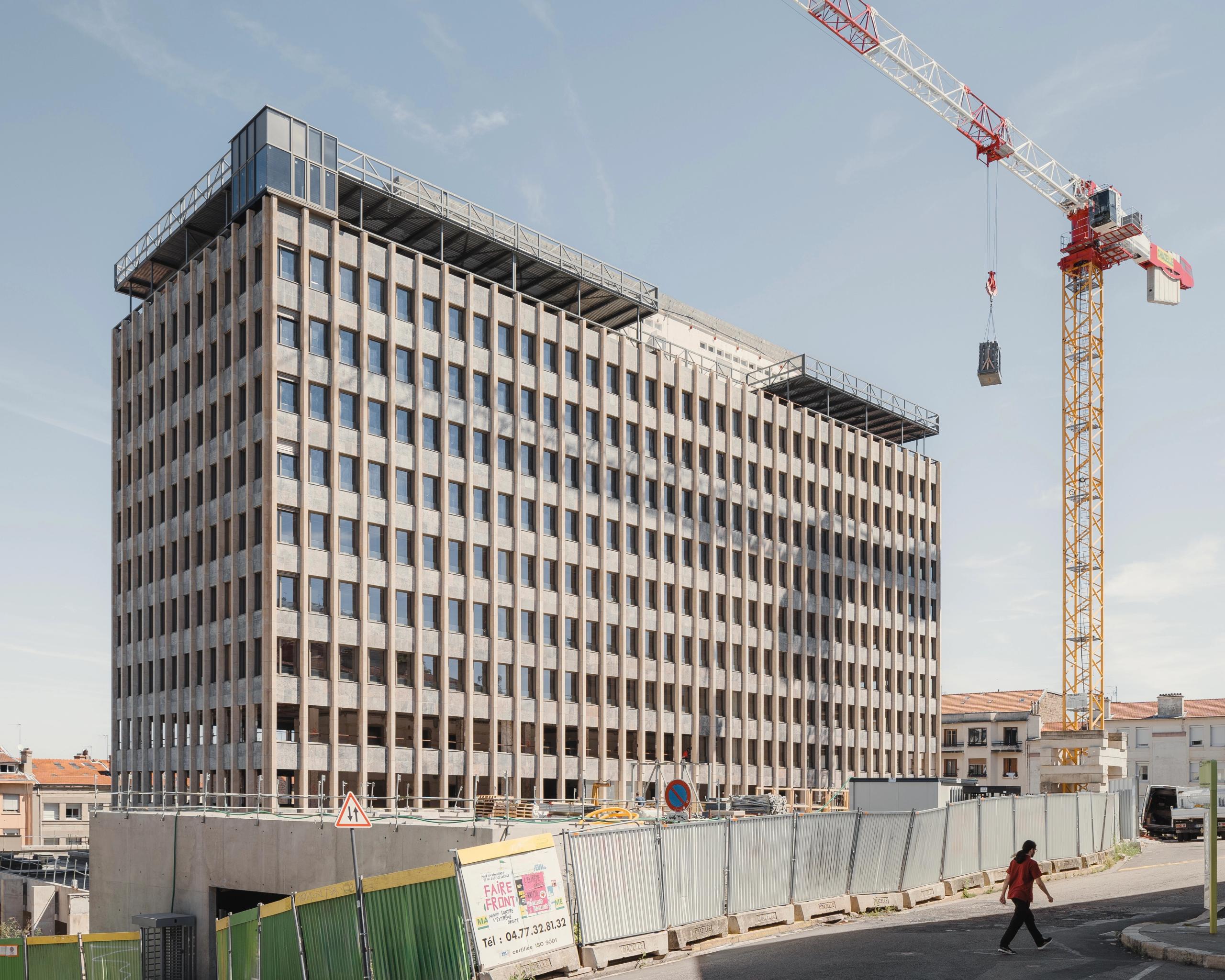 Chantier de réhabilitation de l’IGH Émile Loubet à Saint-Étienne, tour administrative des années 1970 en béton rythmée de trames verticales, grue et clôtures de site.