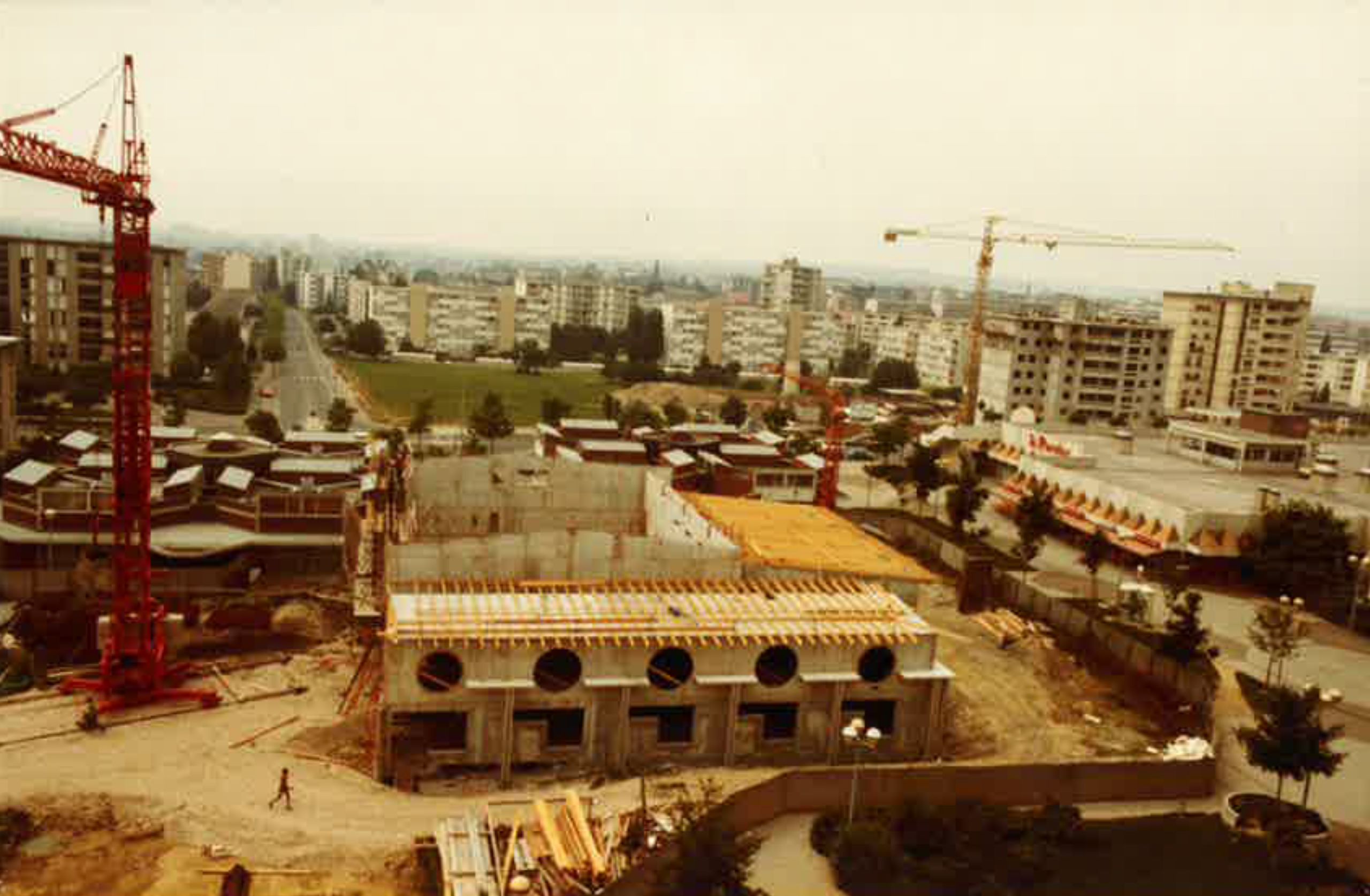 Chantier du conservatoire intercommunal d’Annemasse dans les années 80 : structure en béton, façade percée d’oculi, charpente en cours de pose et grues.
