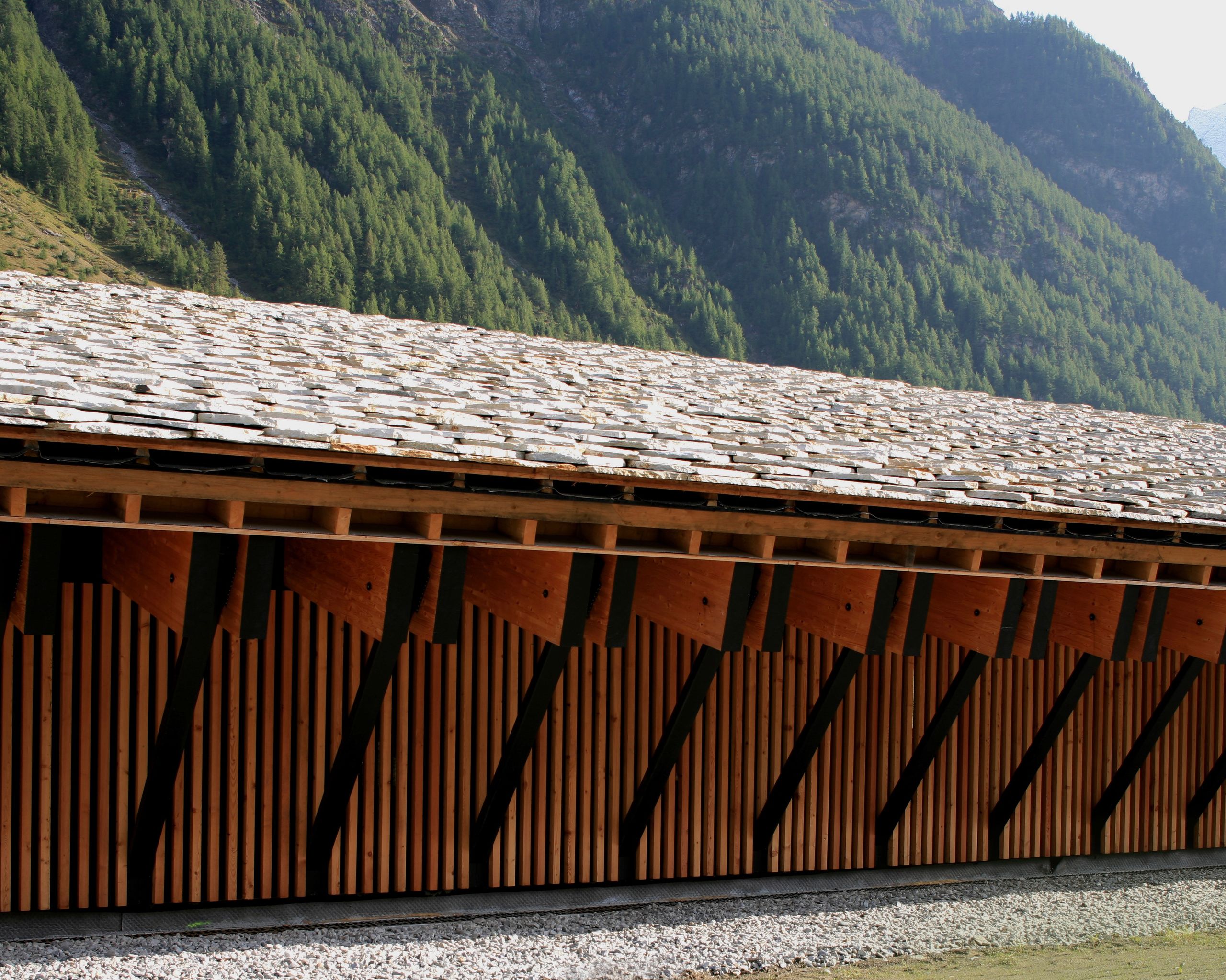 Espace nordique de Bessans : volume linéaire en bois et lauze, façade rythmée de portiques inclinés, inscrit au pied du versant boisé de Haute-Maurienne.