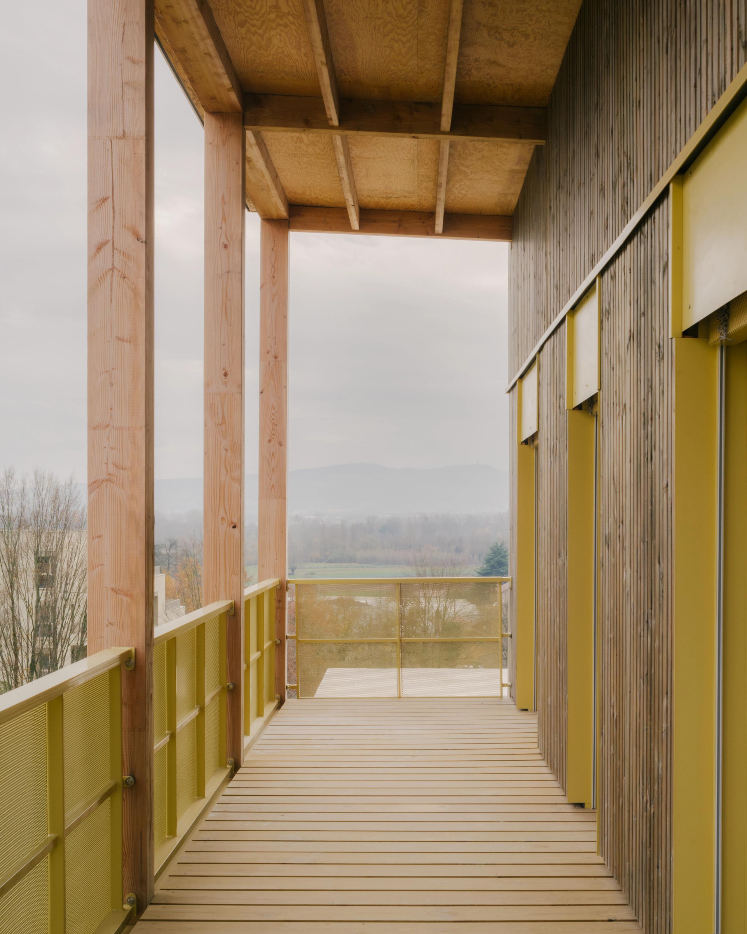 Coursive extérieure en bois et serrurerie jaune des 84 logements à Trévoux, exo-structure poteaux-dalles ouverte sur le parc et le paysage lointain.