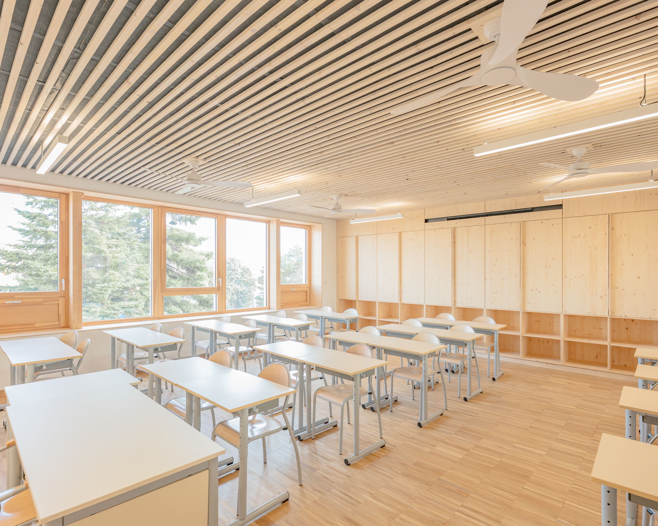 Salle de classe du groupe scolaire J.F. Kennedy à Lyon, structure bois apparente, plafond à lames, grandes baies vitrées et mobilier scolaire aligné.