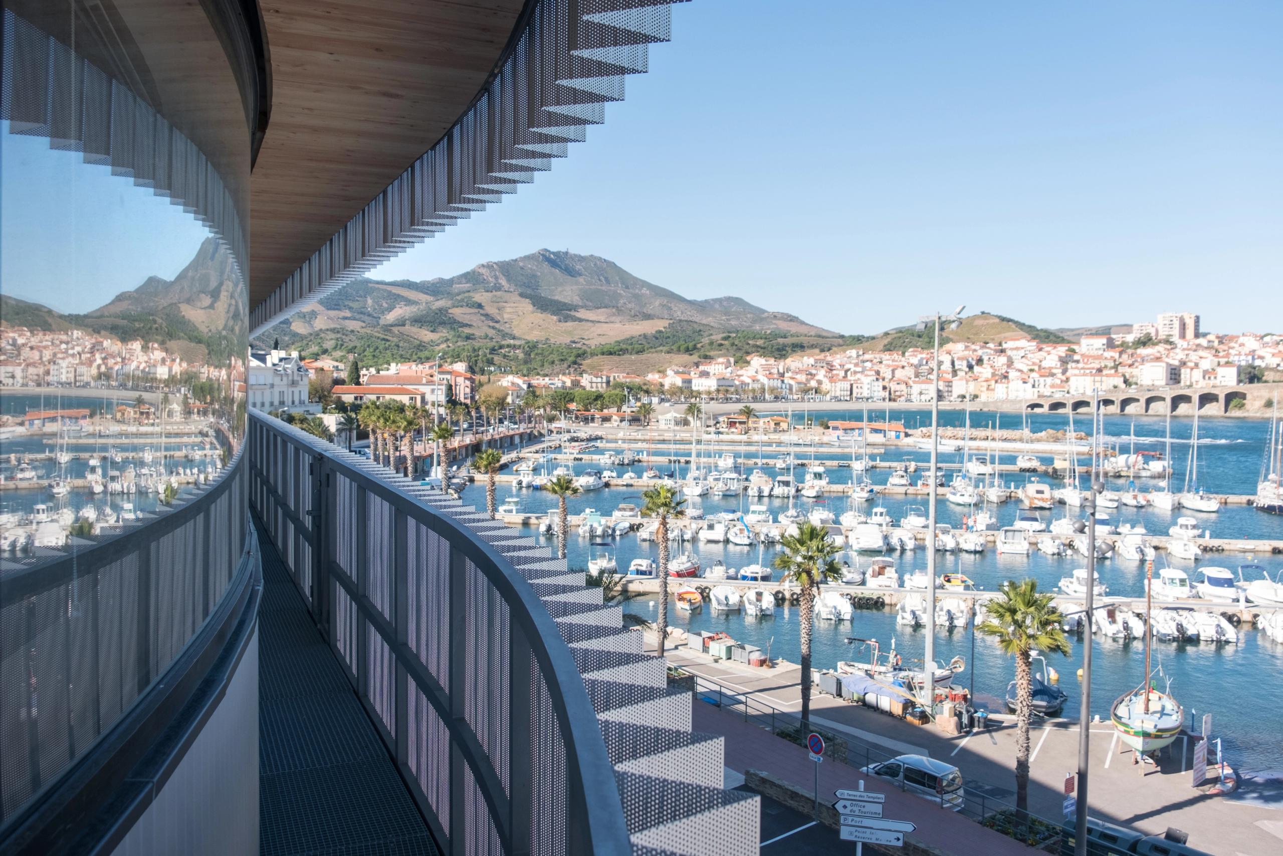 Vue depuis la coursive du Biodiversarium à Banyuls-sur-Mer, façade vitrée courbe et bardage métallique perforé cadrant le port de plaisance et le paysage côtier.