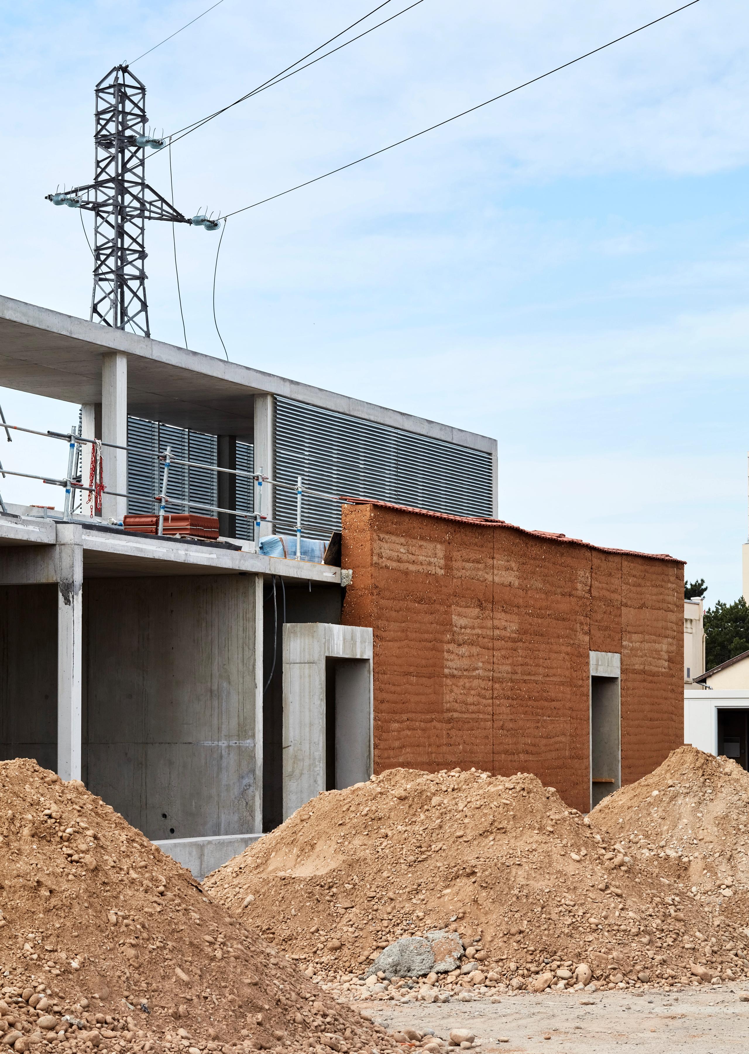 Chantier du poste source de Cusset à Villeurbanne : volumes en béton brut et mur en pisé, structure apparente, terrasse technique et lignes électriques en arrière-plan.