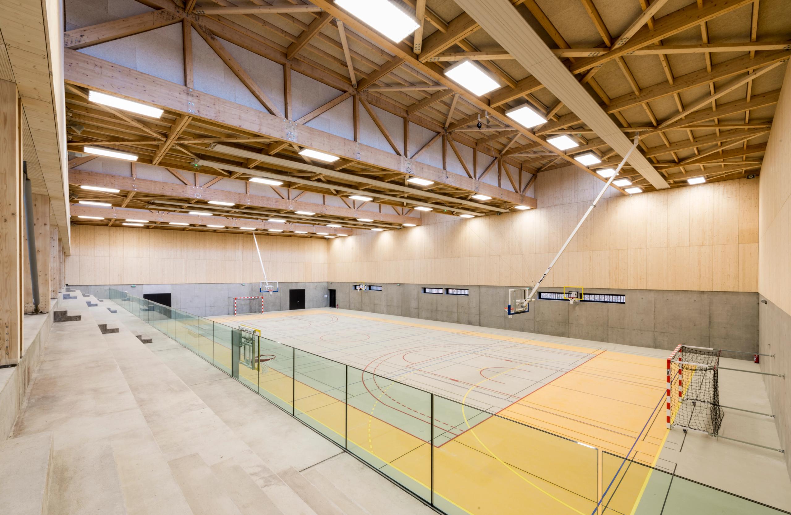 Intérieur du gymnase Hacine Cherifi à Rillieux-la-Pape, grande halle sportive en béton et bois, charpente treillis apparente et gradins minéraux latéraux.