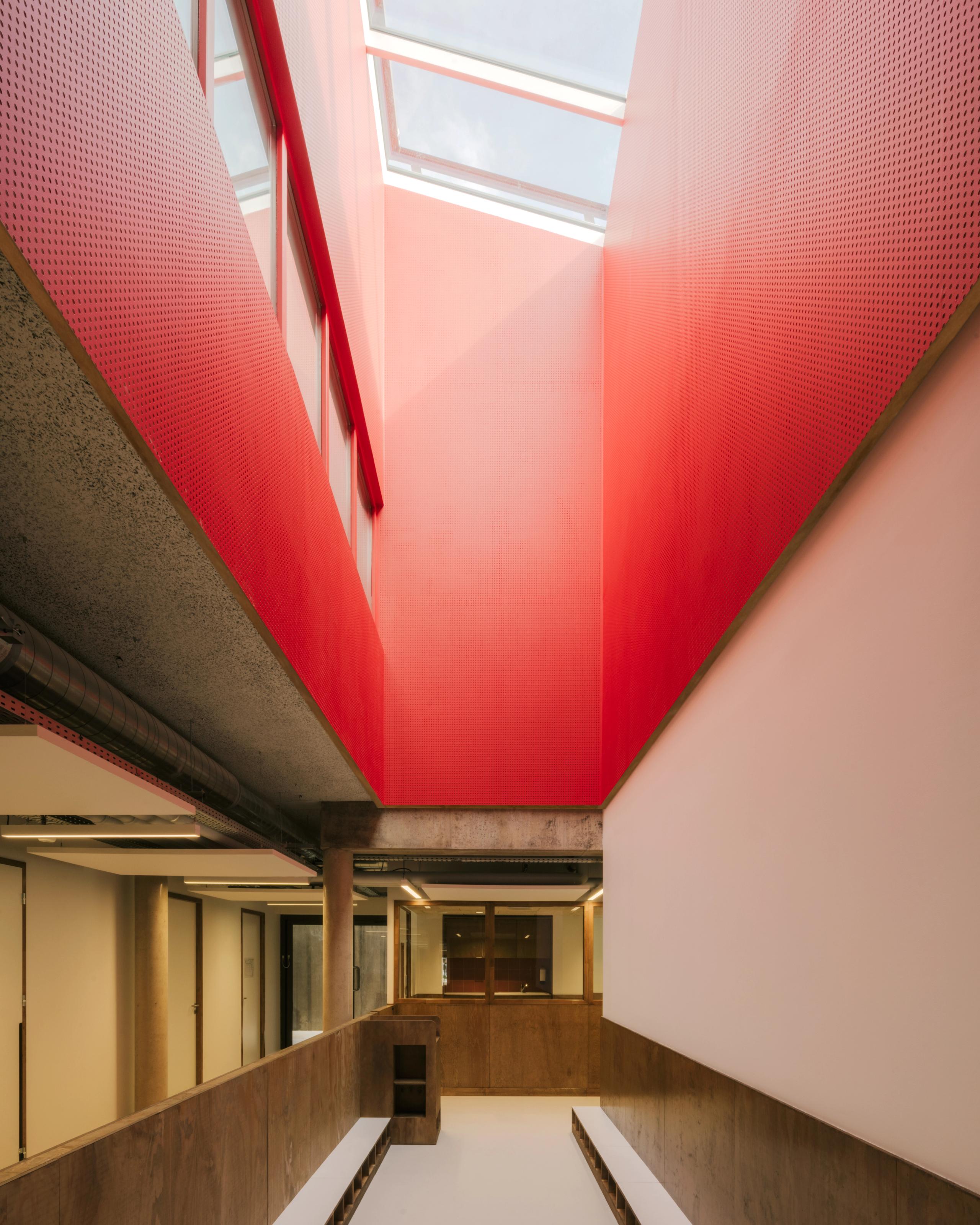 Atrium de la Maison des familles à Limonest, circulations en double hauteur sous puits de lumière, parois rouges perforées et structure béton-bois apparente.