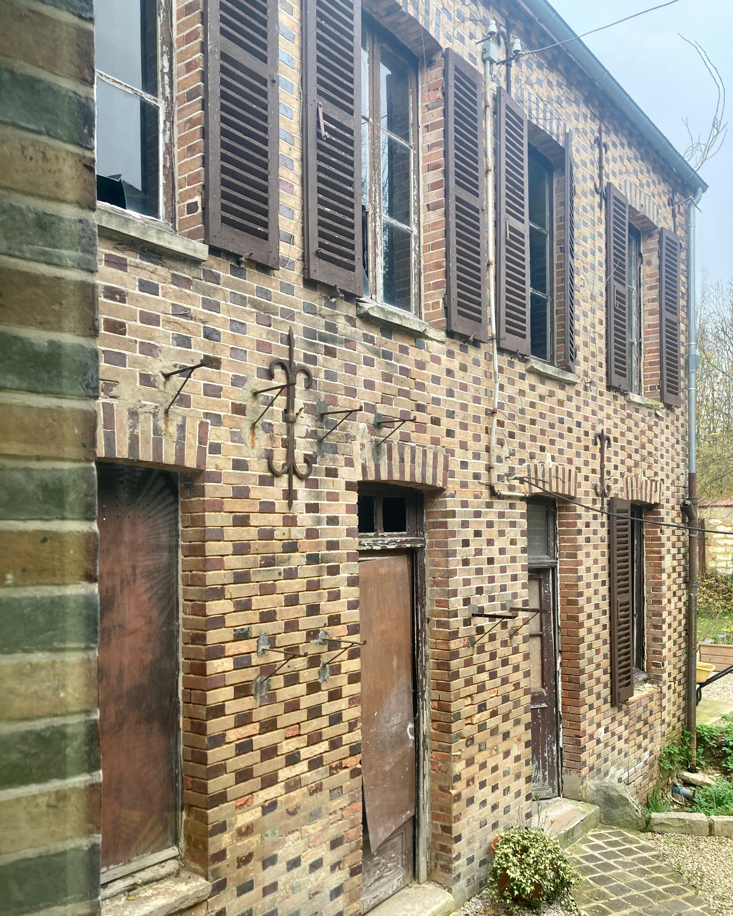 Façade en brique d’un ancien bâtiment à volets bois et menuiseries dégradées, illustrant l’état existant avant intervention à Saint-Valérien.