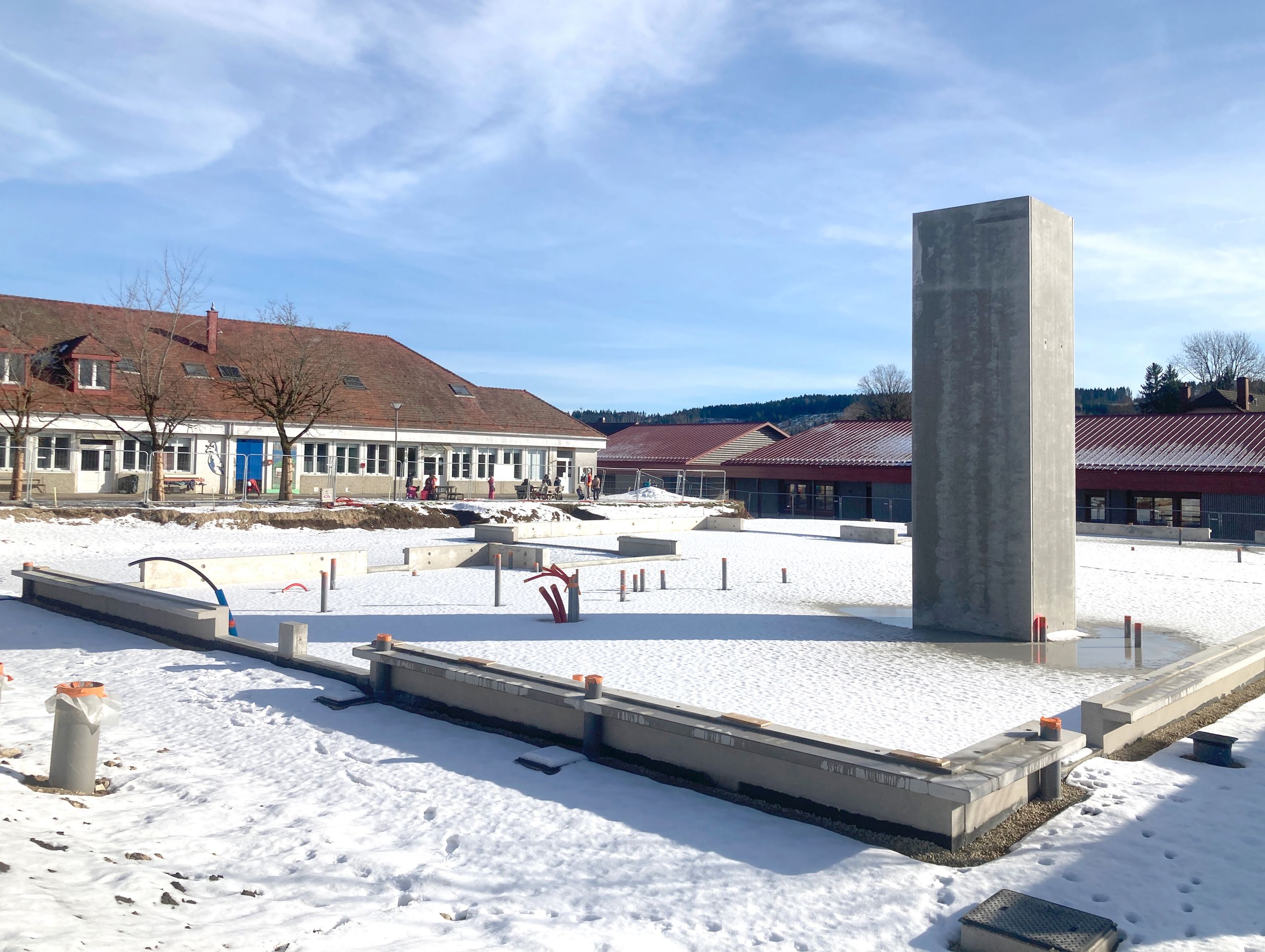 Chantier enneigé du School complex Louis Pergaud à Levier, montrant les fondations en béton, un noyau vertical et les bâtiments scolaires existants en arrière-plan.