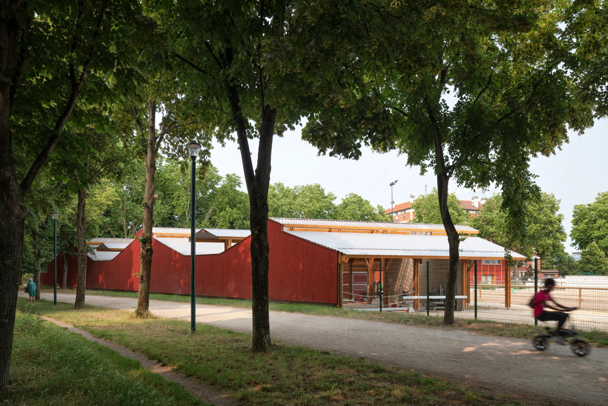 Street-side view of Tectoniques’ Falun-red timber equestrian center in Gennevilliers, with sawtooth roofs lining a tree-shaded path beside the arenas.