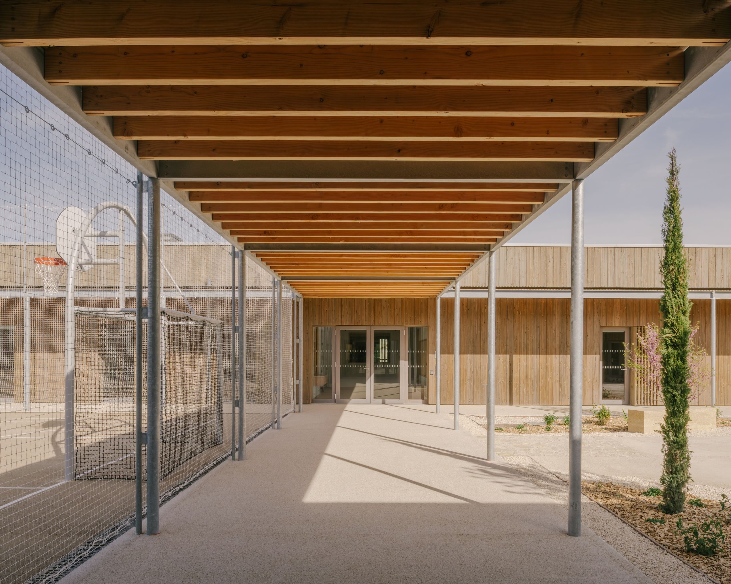 Passage couvert du pôle culture et jeunesse de Vaison-la-Romaine, structure bois-métal cadrant la cour minérale entre façades bardées de bois et jardin planté.