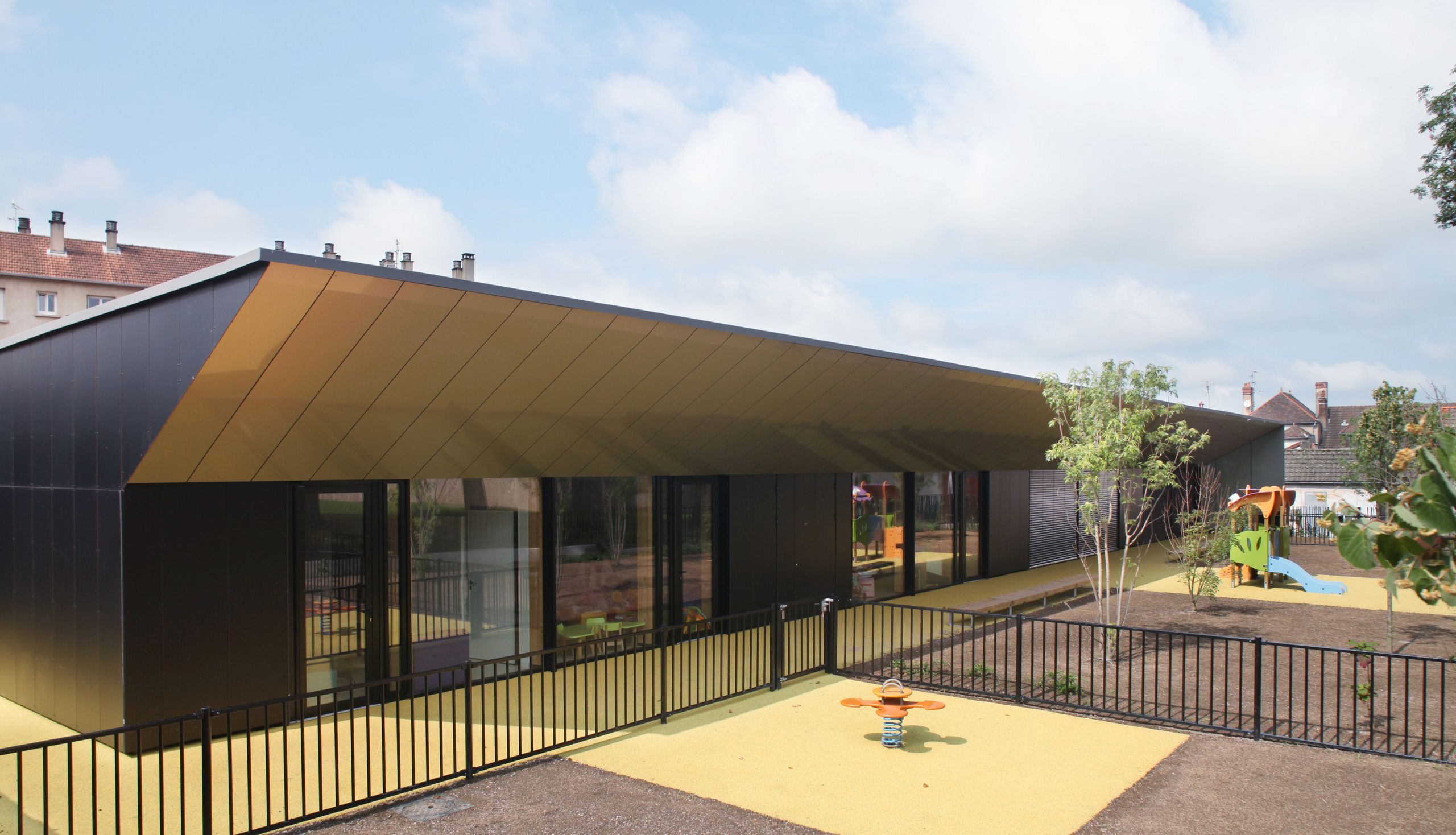 Maison de la petite enfance à Autun : volume bas en équerre, façades sombres vitrées sous large casquette métallique dorée, donnant sur cour et aire de jeux.