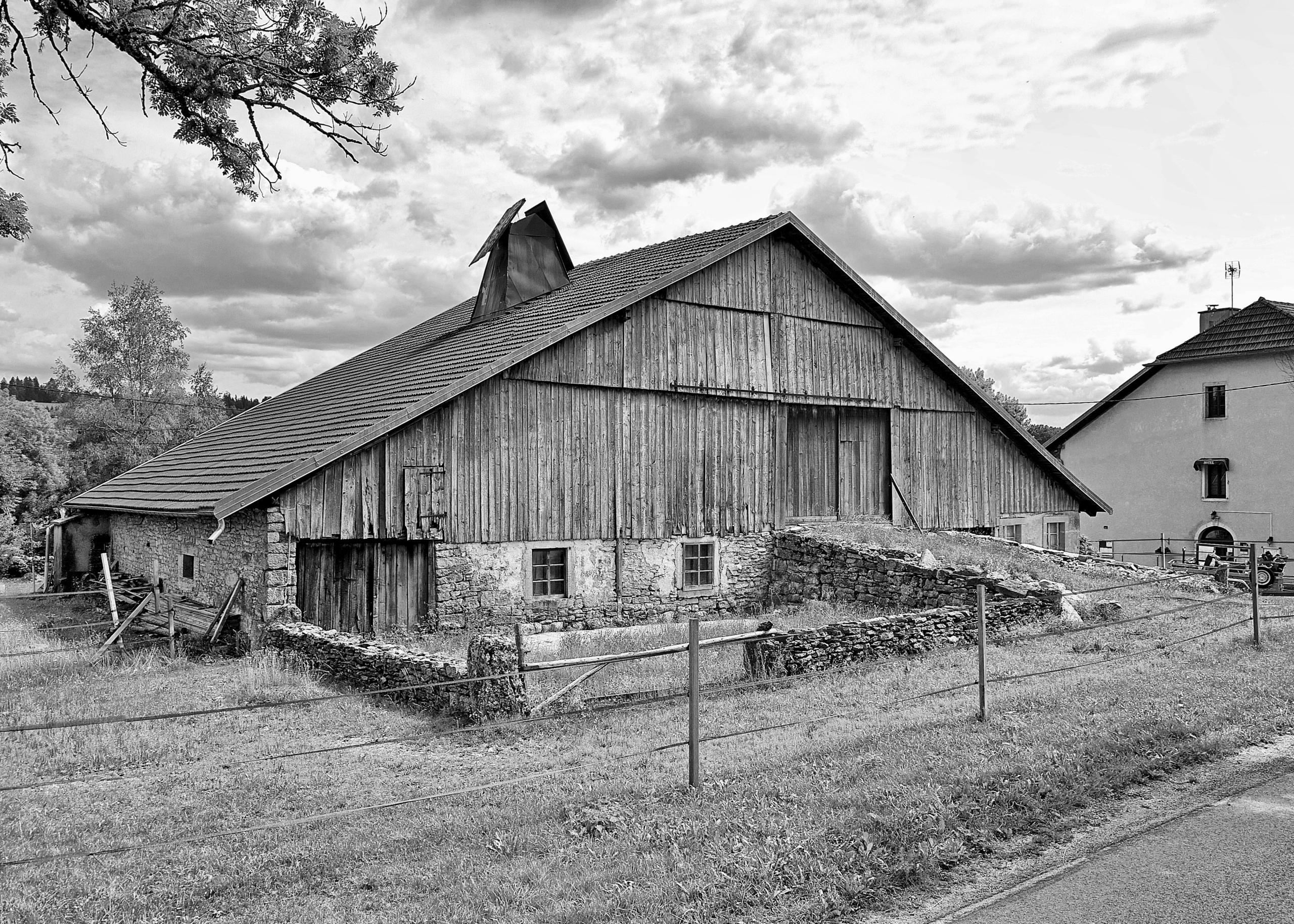 Bâtiment rural vernaculaire à Levier, volume longiligne en pierre et bardage bois sous grande toiture à deux pans, contexte forestier du futur groupe scolaire Louis Pergaud.