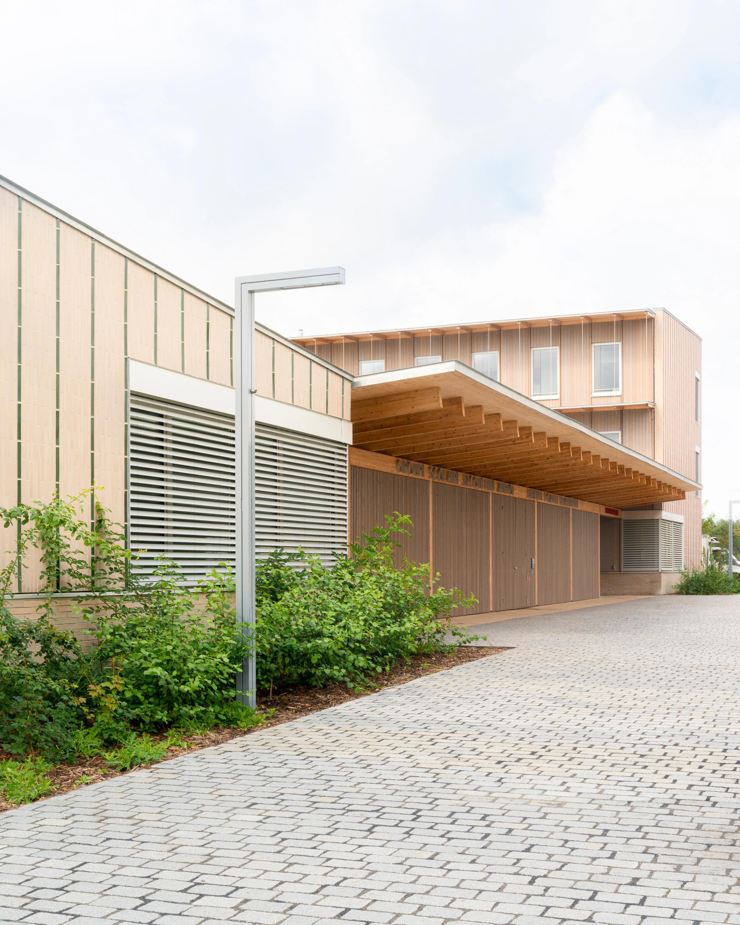 Façade bois et terre cuite des groupes scolaires Jacqueline Auriol et Jean Jaurès au Bourget, préau couvert en saillie et cheminement minéral planté.