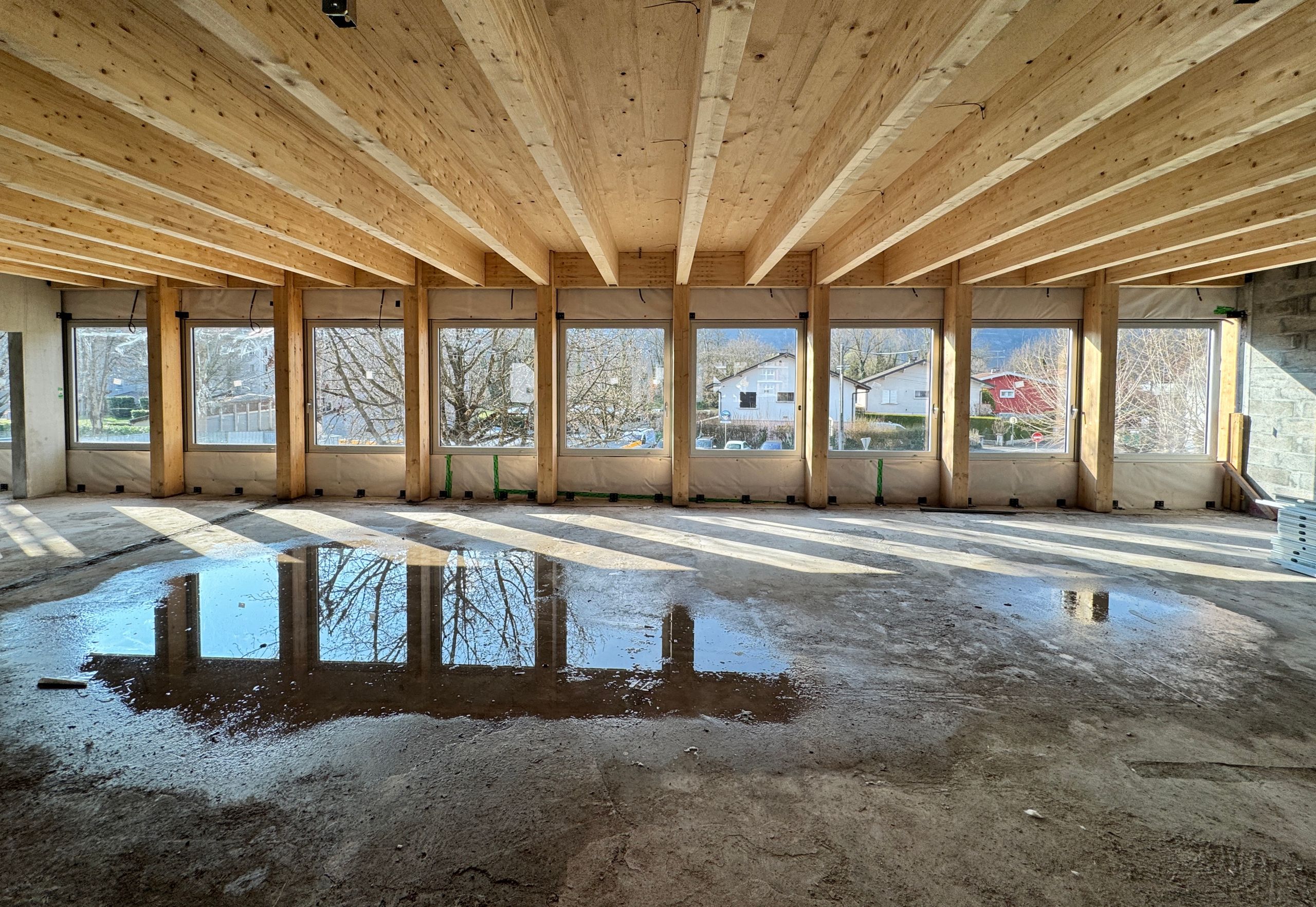 Intérieur en chantier du groupe scolaire du Bouchet à Bonneville, grande salle en ossature bois avec baies vitrées régulières et dalle brute réfléchissante.
