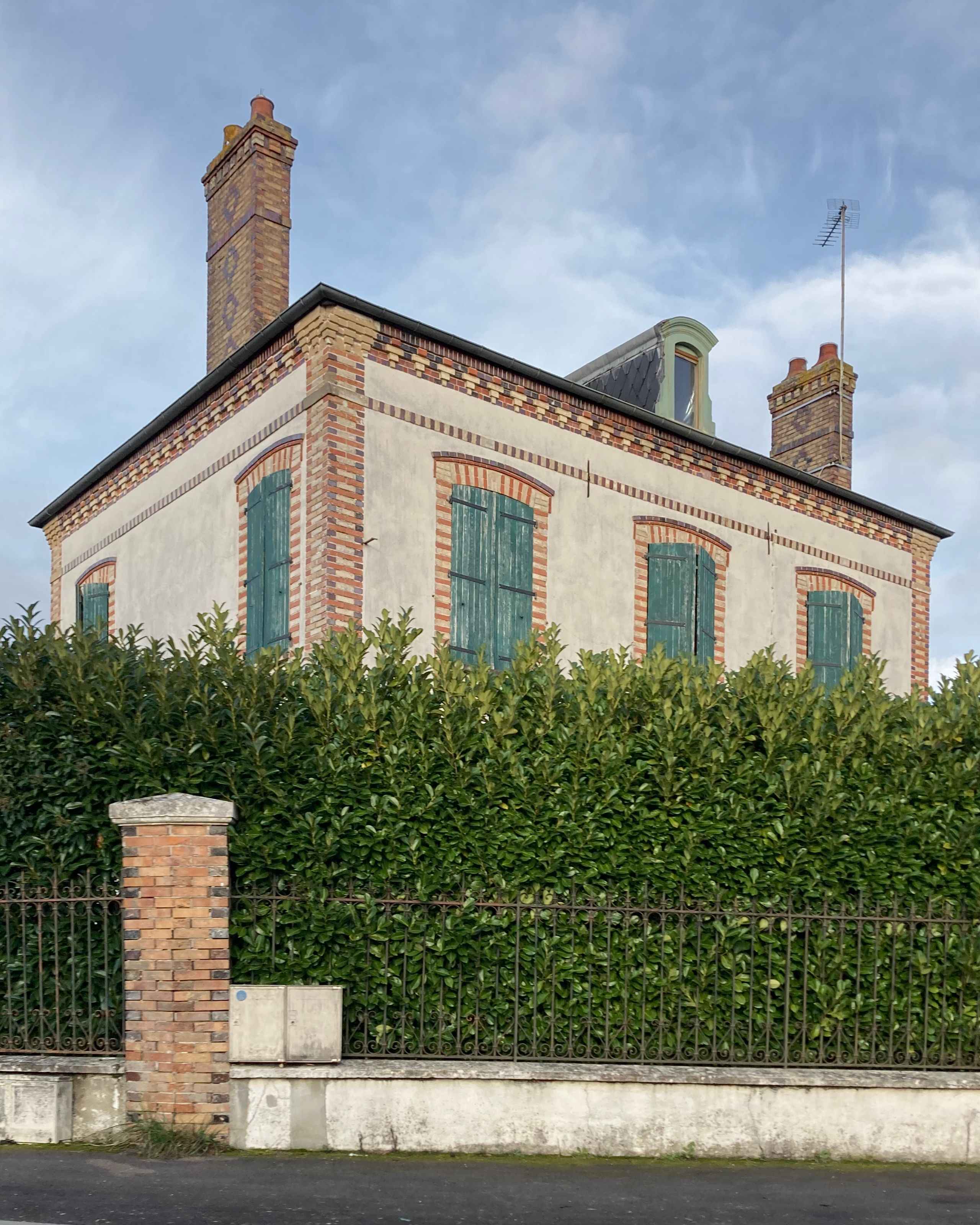 Maison en briques enduites à volets bois verts, toiture ardoise et souches de cheminée, vue depuis la rue derrière une haie dense et une clôture métallique.