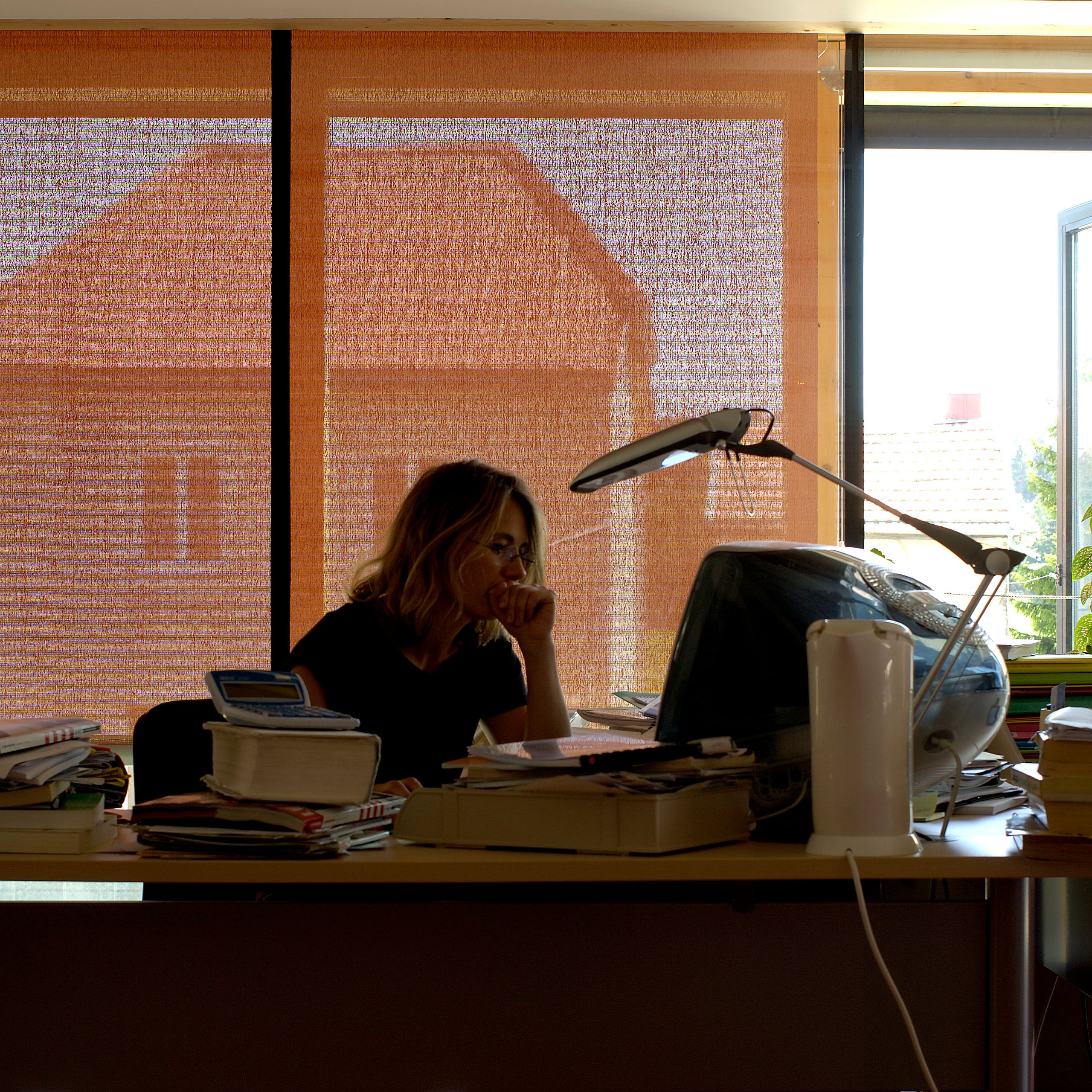 Bureau d’architecte Tectoniques à Lajoux, poste de travail face à une grande baie vitrée filtrée par stores, vue diffuse sur les toitures du village.