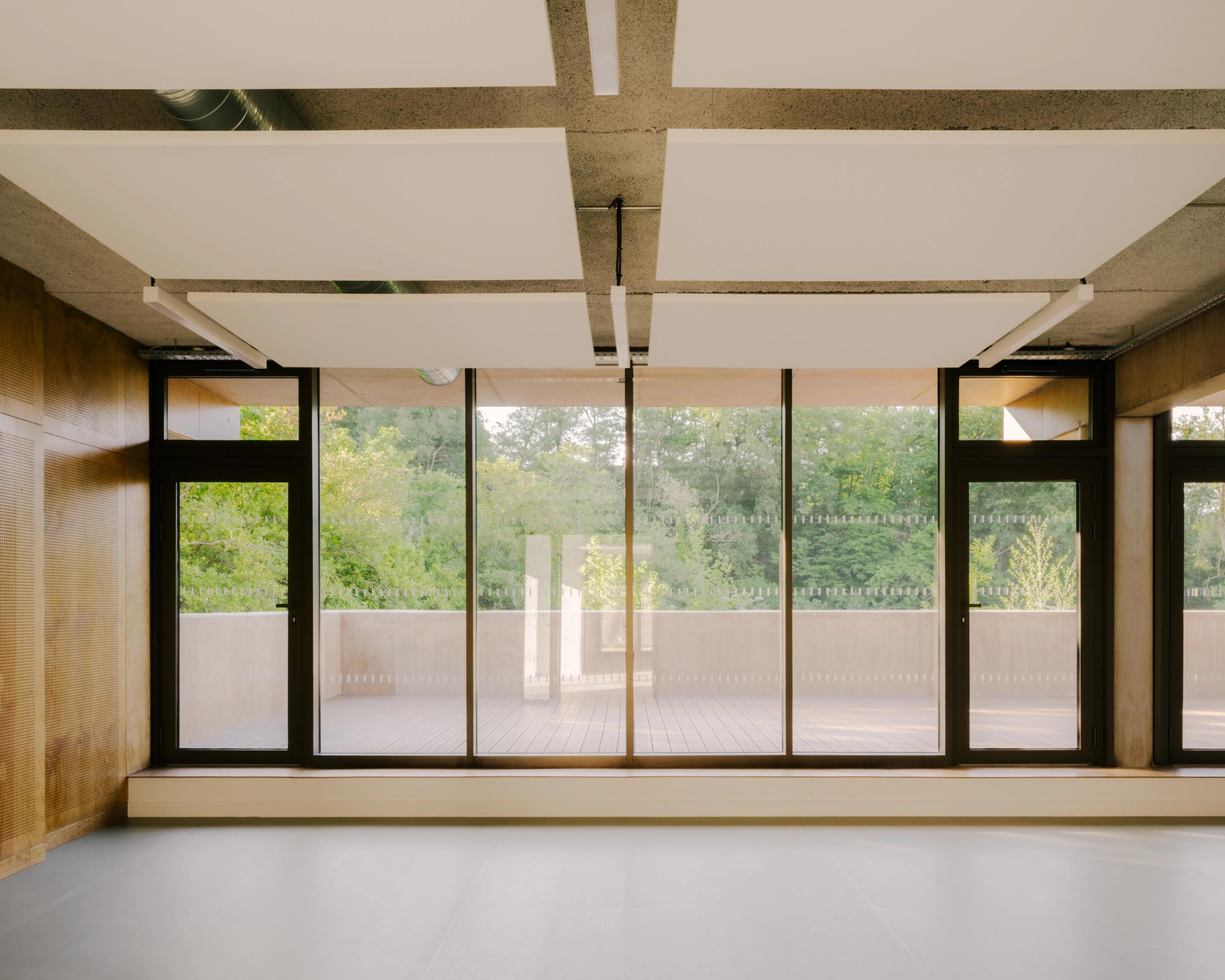 Intérieur de la Maison des familles à Limonest : plateau libre en béton, parois bois, plafond technique avec panneaux acoustiques et large façade vitrée sur terrasse paysagère.