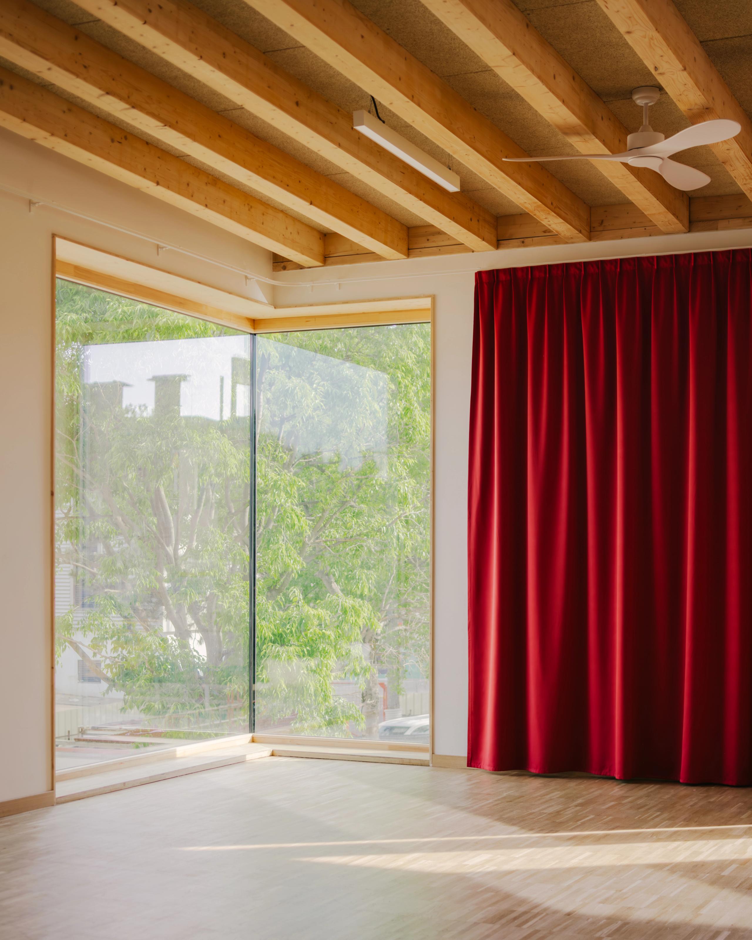 Intérieur du groupe scolaire Kennedy à Lyon : salle lumineuse en structure bois, grande baie vitrée d’angle sur la cour et rideau acoustique rouge.