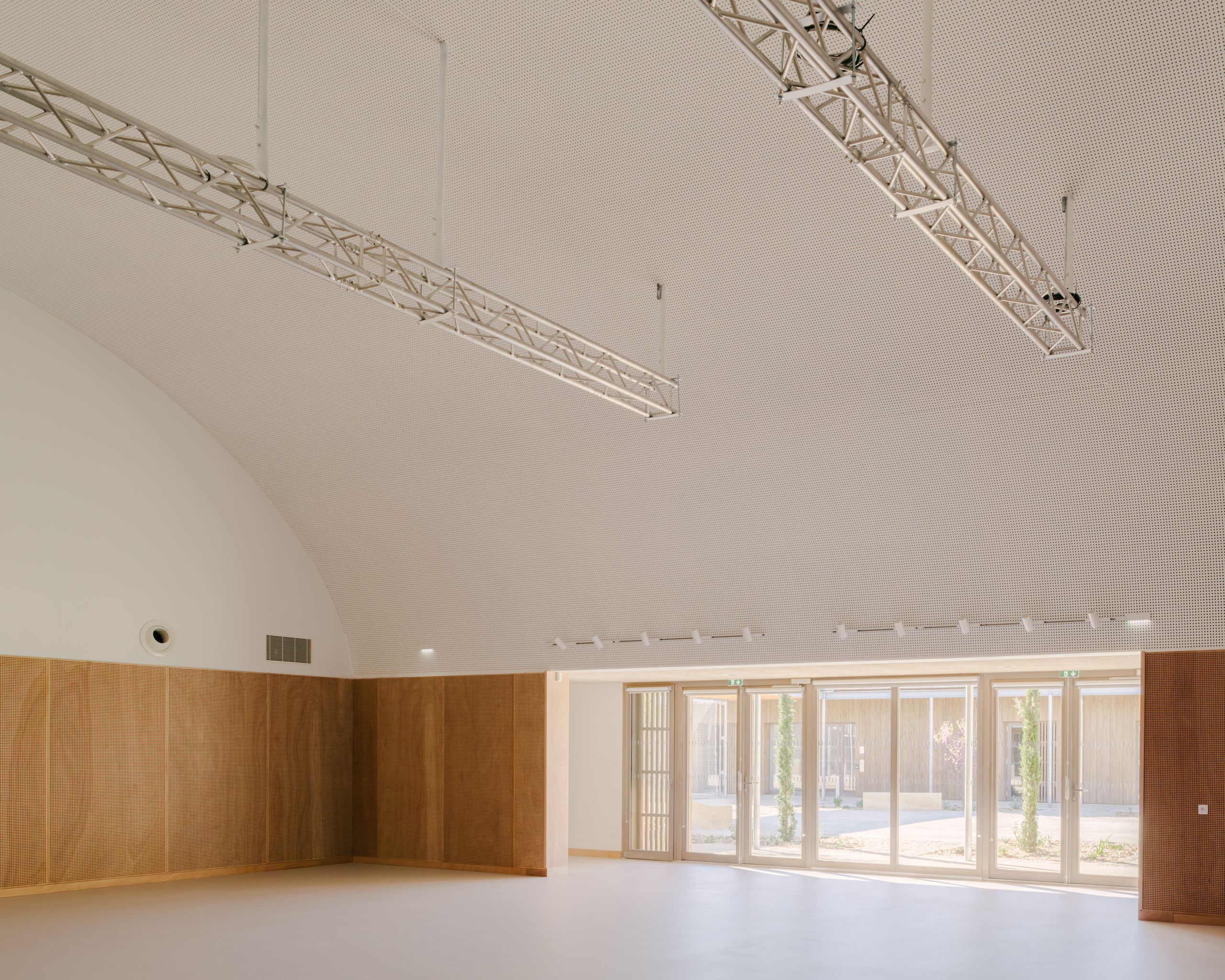 Grande salle du pôle culture et jeunesse à Vaison-la-Romaine, voûte acoustique blanche, panneaux bois perforés et façade vitrée ouvrant sur le patio planté.